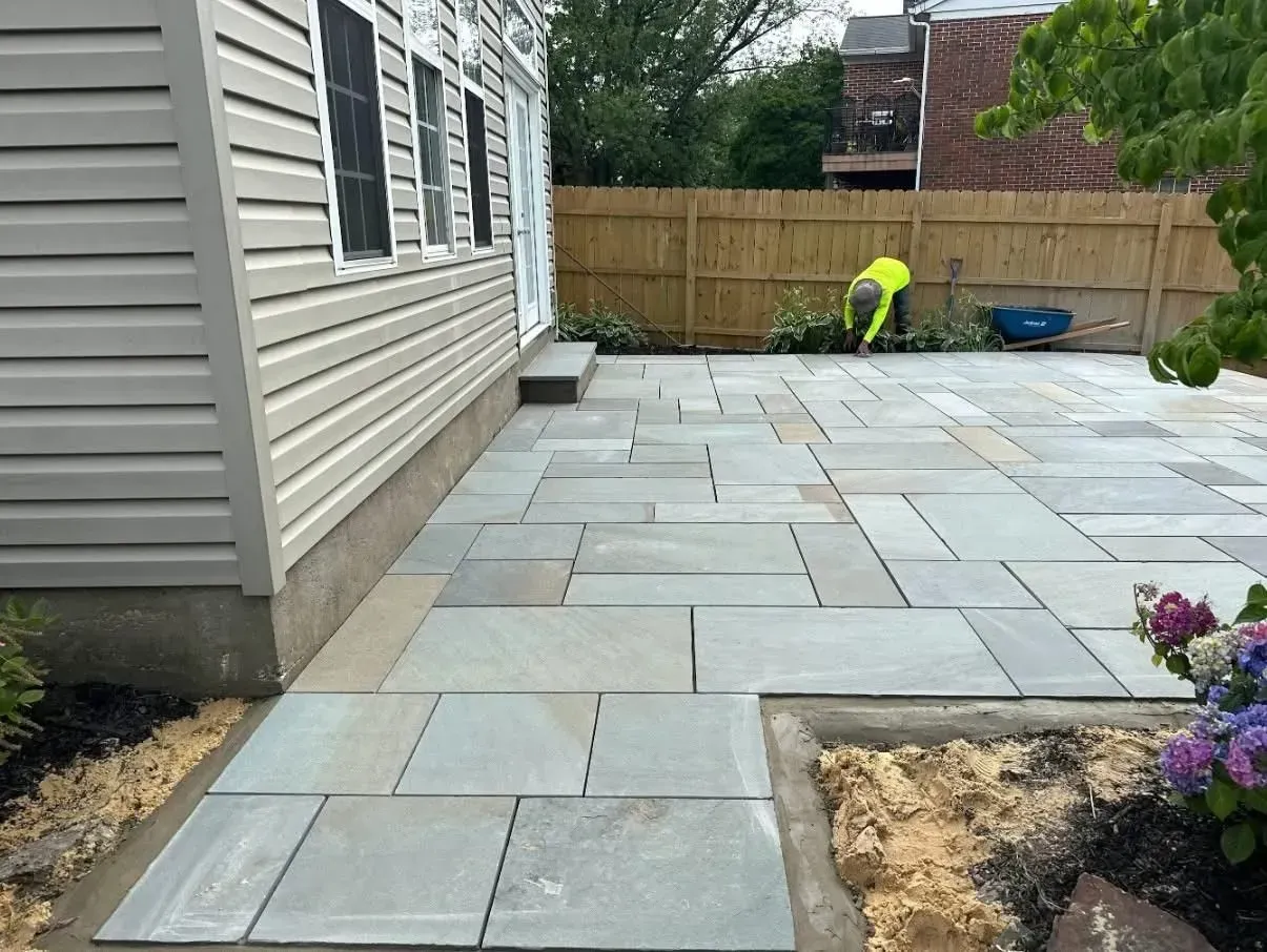 Newly constructed outdoor patio with rectangular gray stone pavers next to a light-colored house and a wooden fence. A worker is in the background.