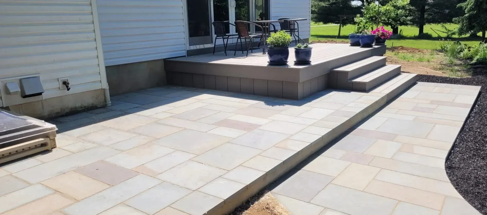 A paved patio with steps leading to a raised deck area with potted plants, near a house with siding and an open door, surrounded by a green lawn.