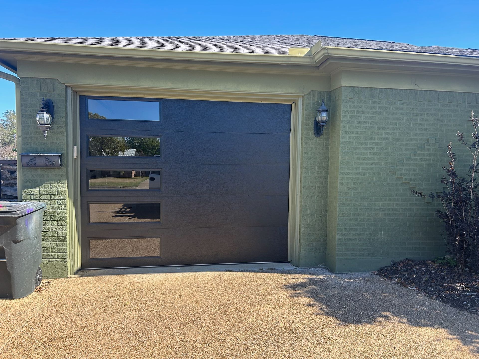 A green house with a black garage door and a trash can in front of it