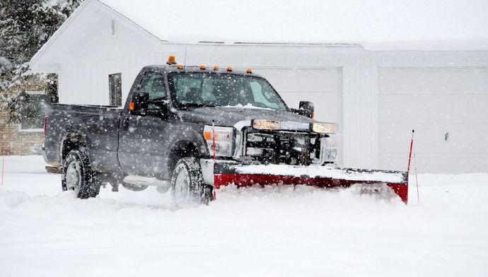 Pickup truck with red snow plow clearing deep snow in a snowy driveway by a house.