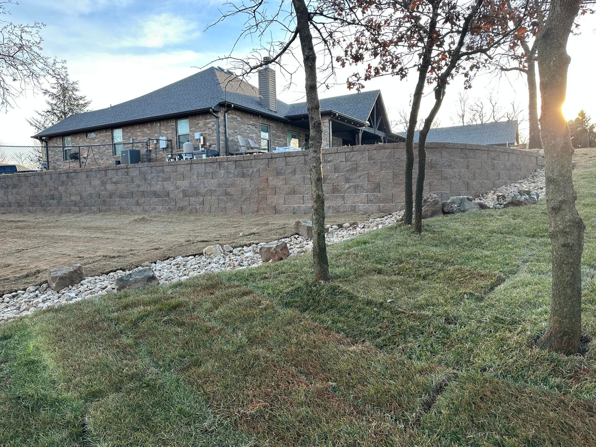Stone house behind a low retaining wall, with grassy foreground and bare trees at sunset.