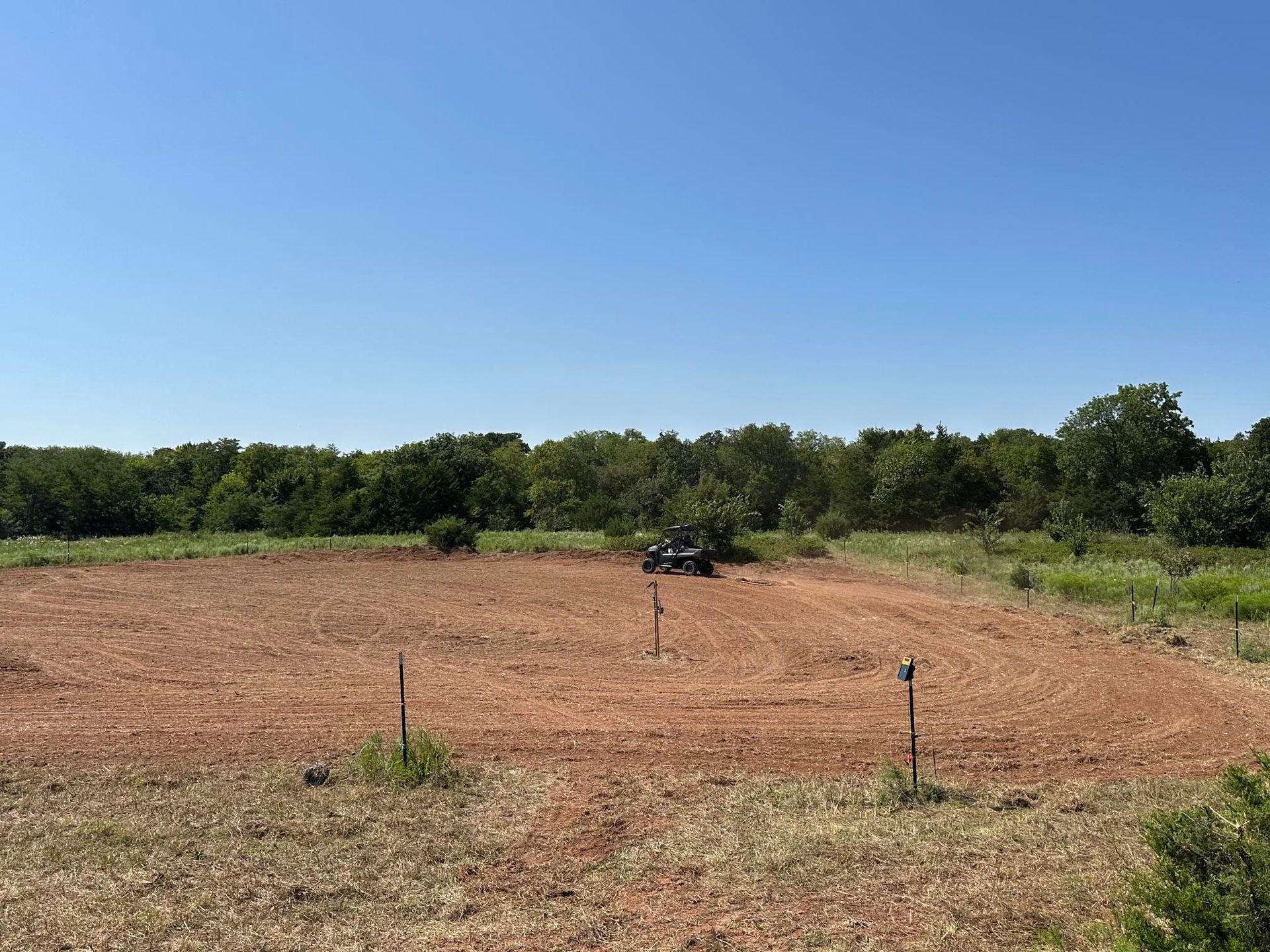 Open field with a lone vehicle and small posts under a clear blue sky, bordered by trees