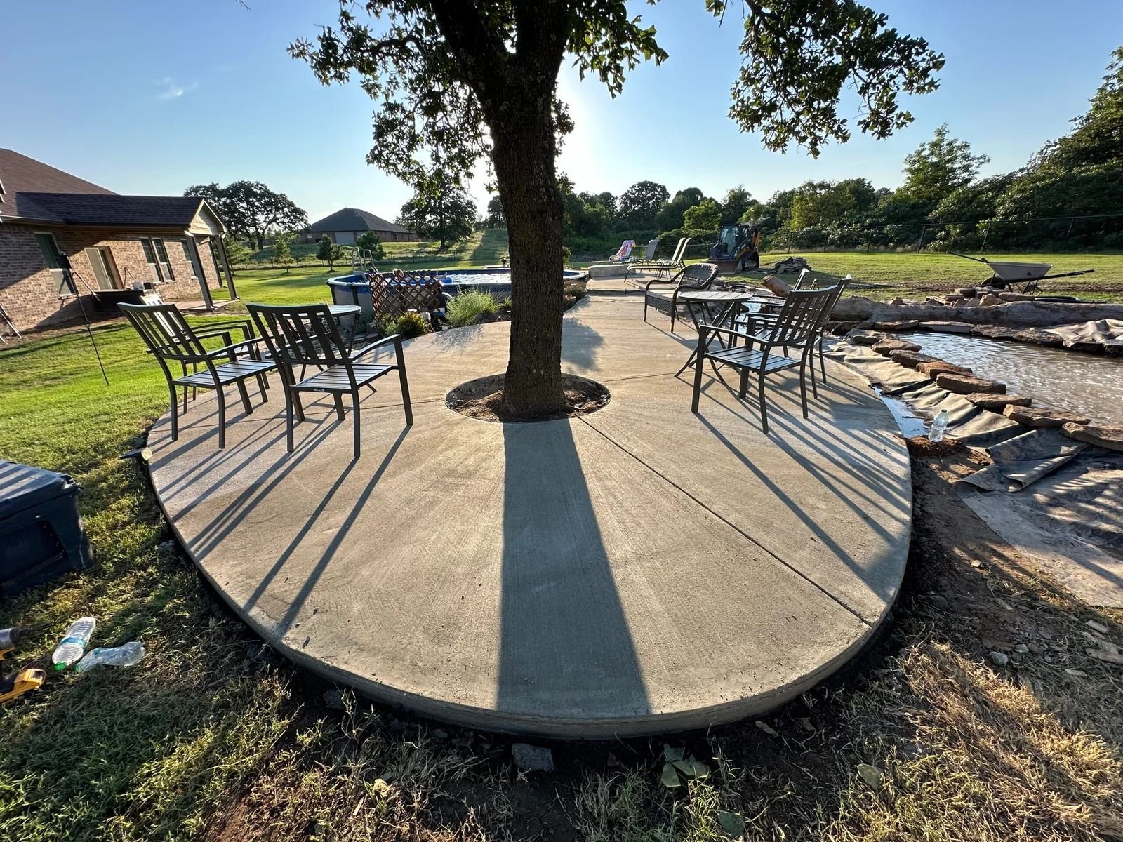 Round concrete patio with metal chairs and a central tree in a sunny yard