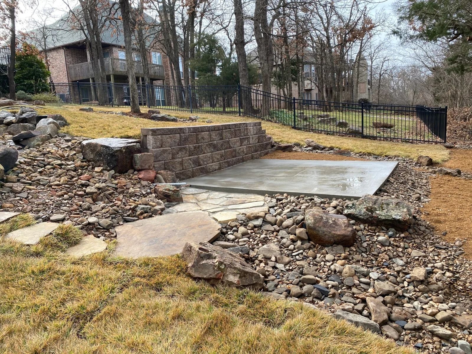 Stone retaining wall and concrete patio in a sloped backyard with rocks and bare trees.