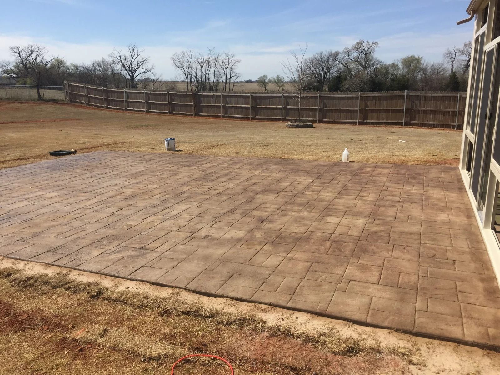 Empty brick patio in a fenced backyard with dry grass and trees under a clear sky
