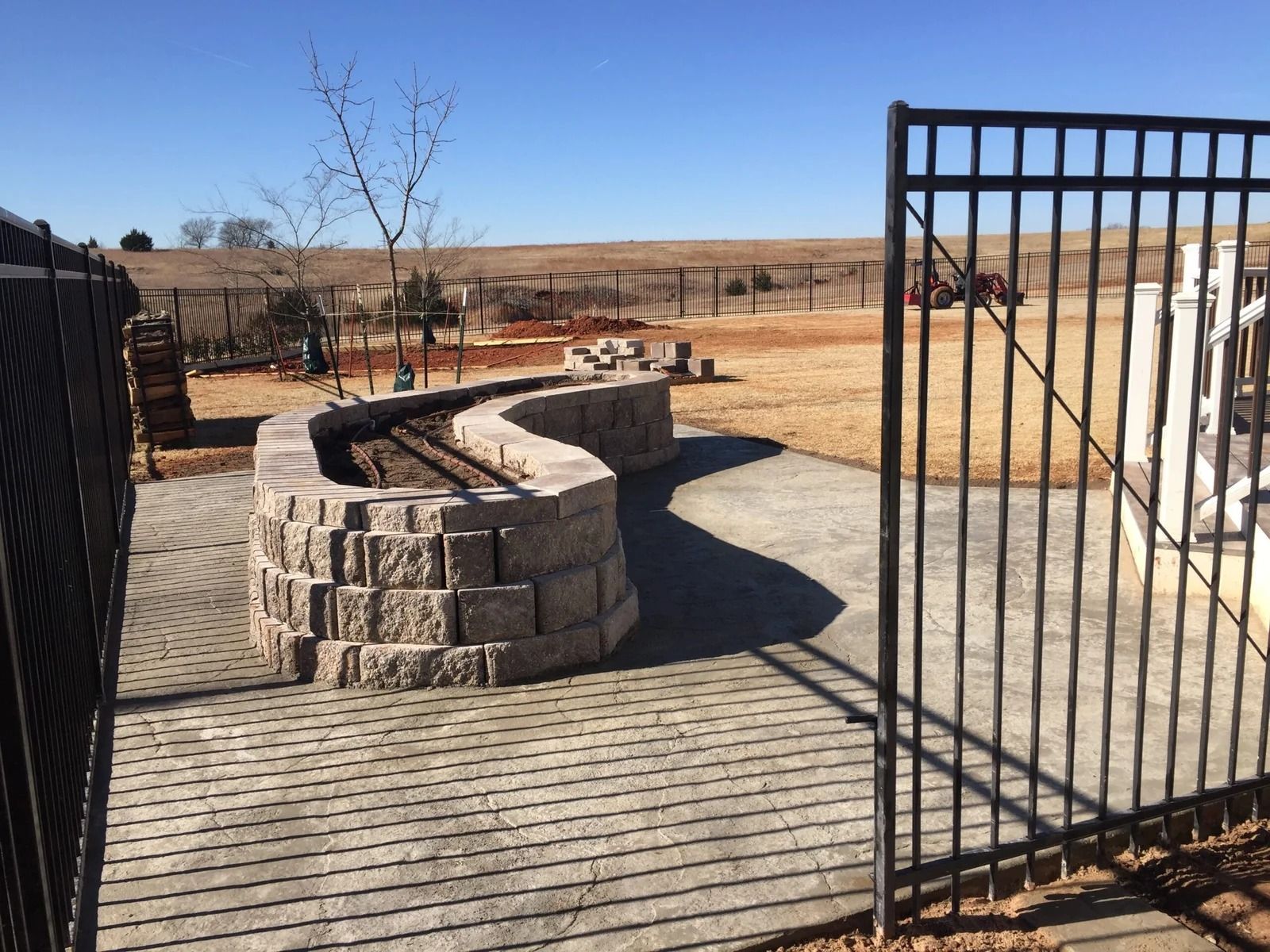 Stone fire pit and grill area on a wooden deck, viewed through an open black gate in a dry field.