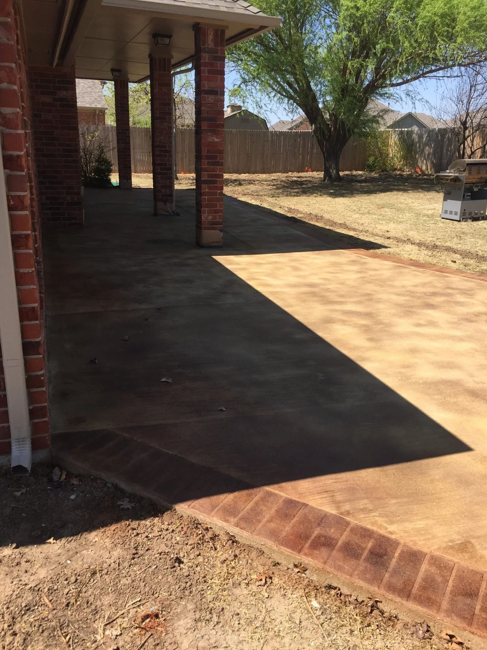 Shaded brick porch with concrete floor and steps, opening onto a sunny yard with a tree and fence.