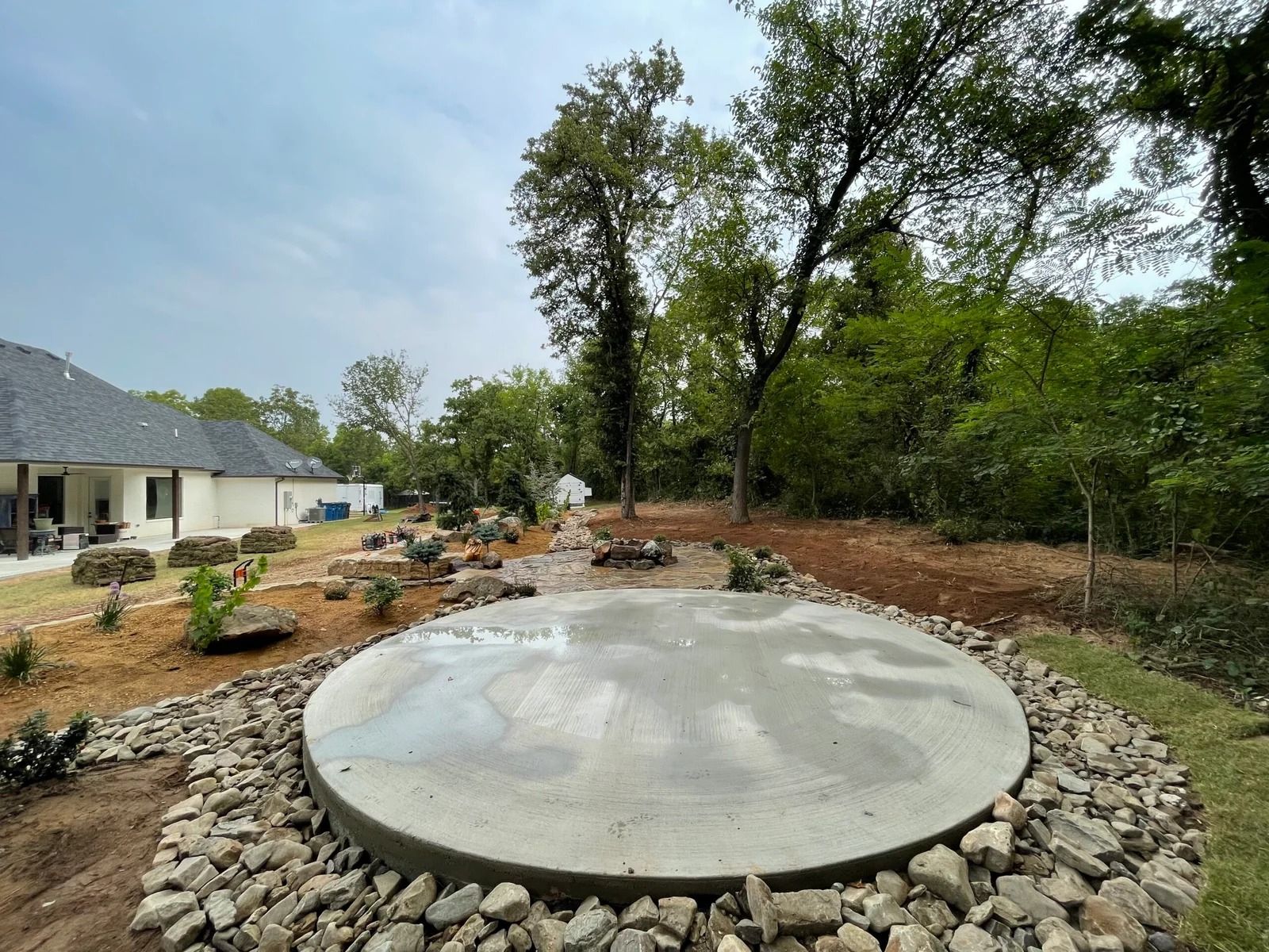 Circular concrete fire pit in a landscaped backyard with rocks, trees, and a house in the background
