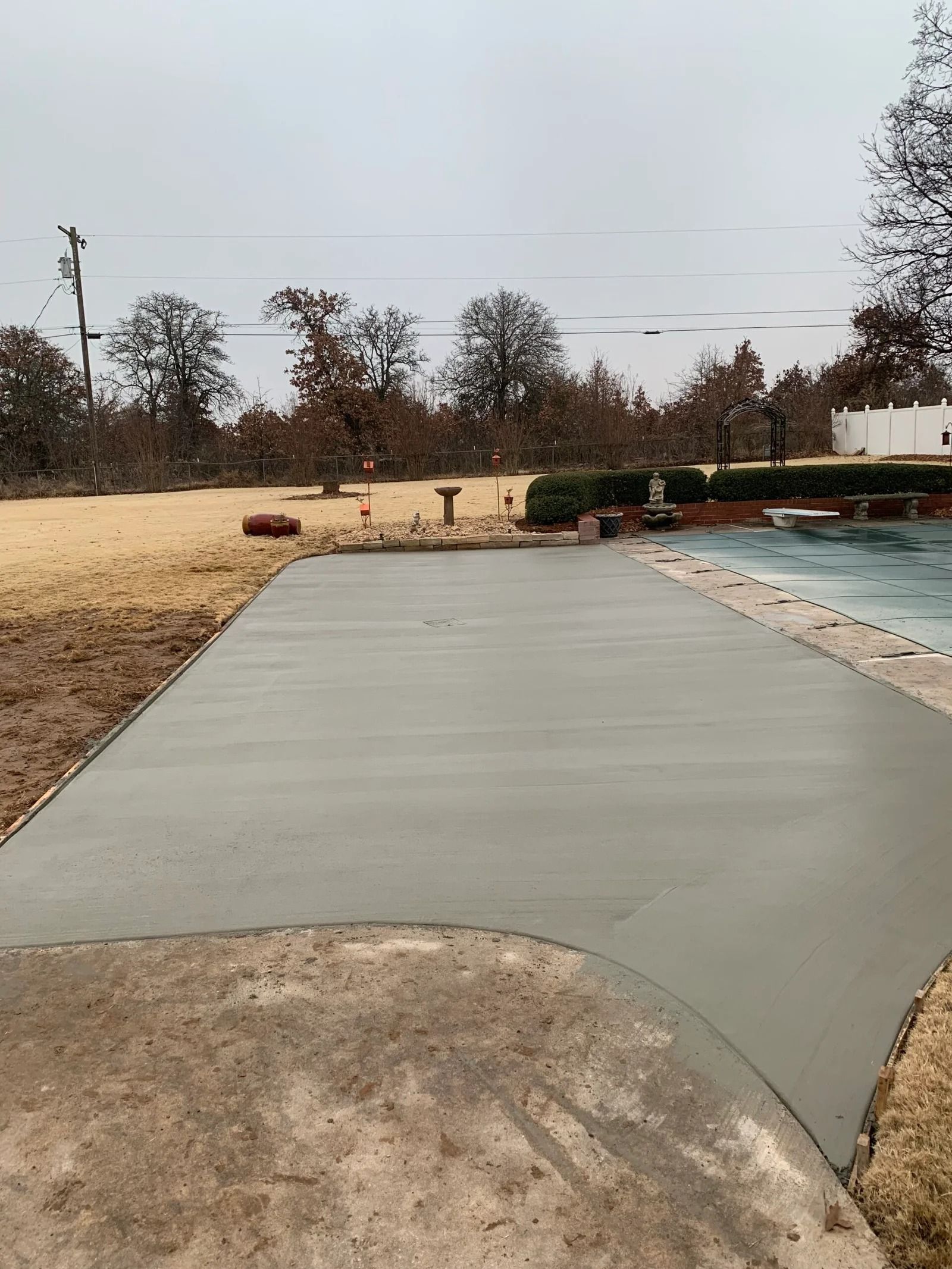 New concrete walkway beside a pool, bordered by winter grass and trees under an overcast sky