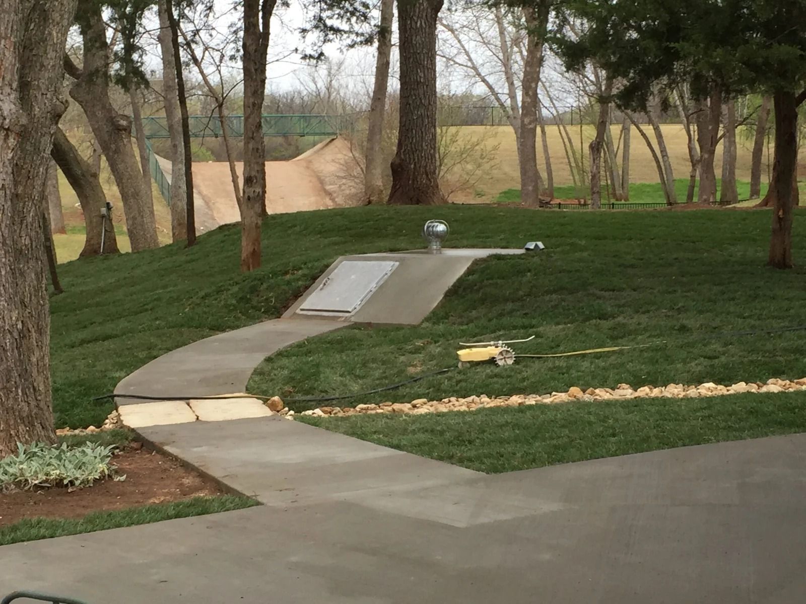 Curved concrete walkway through a grassy yard with trees and a small ramp, leading toward a field.