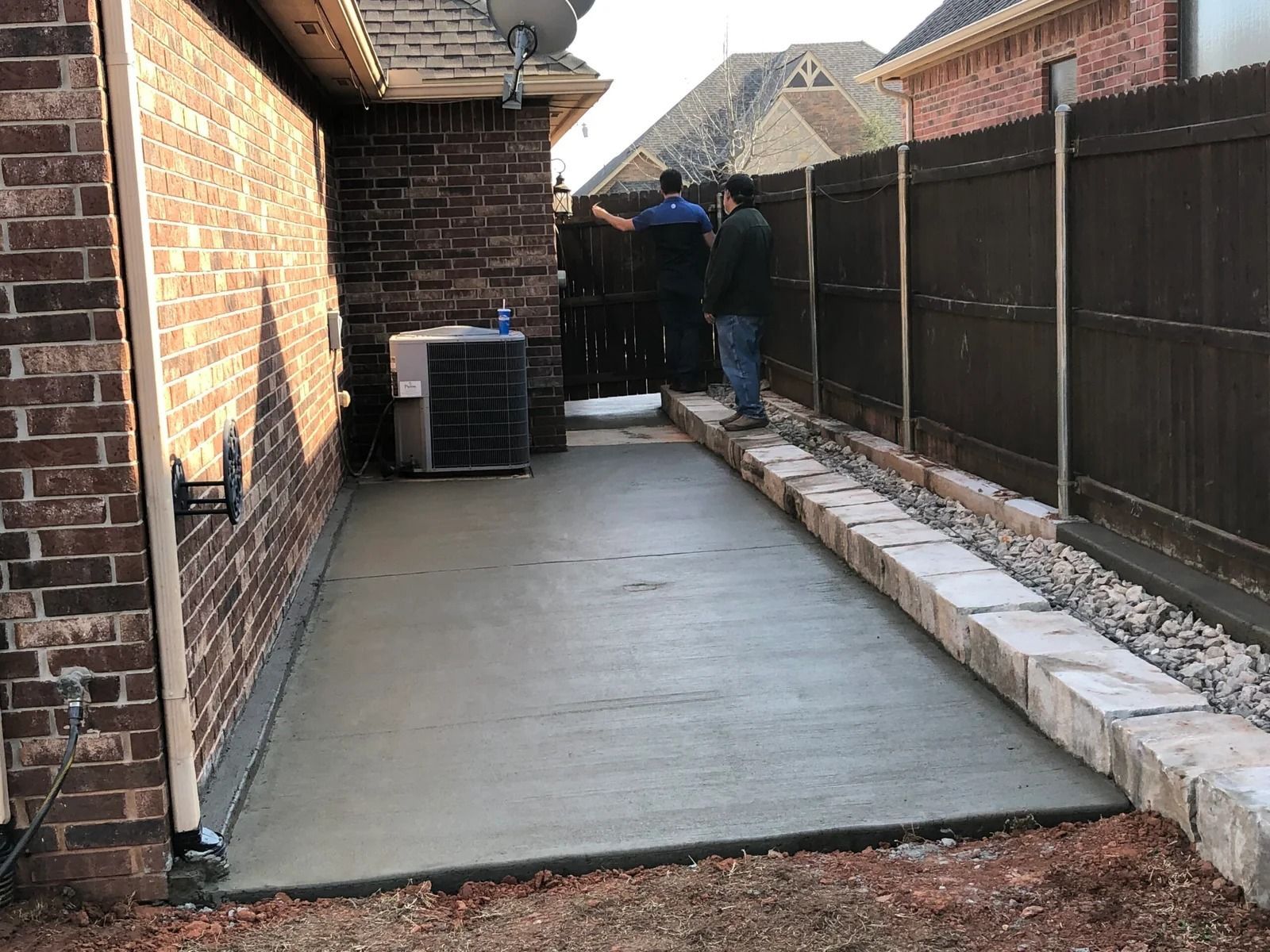 Backyard concrete patio with two workers near a brick house and a gravel border