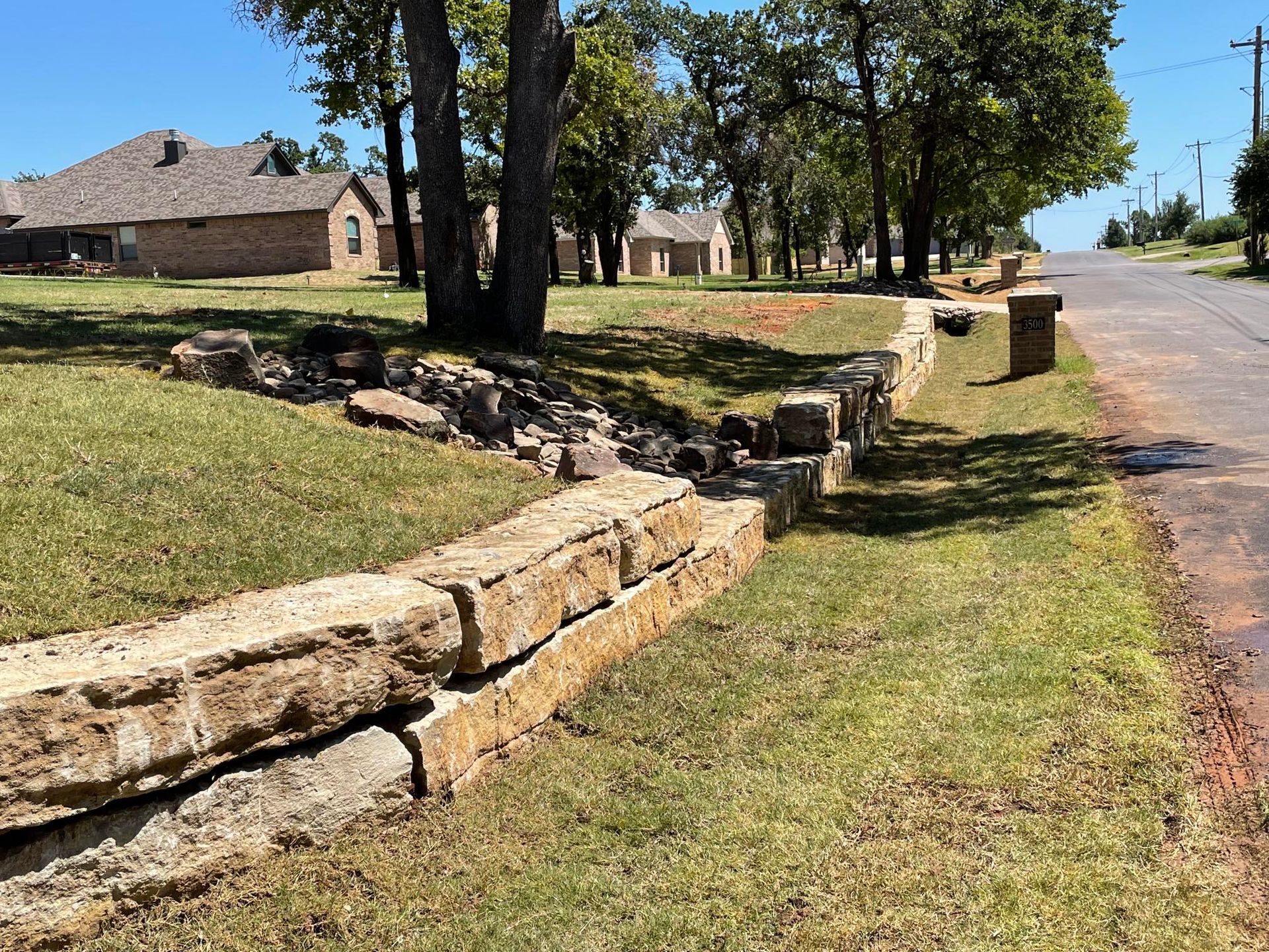 Stone retaining wall along a grassy roadside with houses and trees in the background
