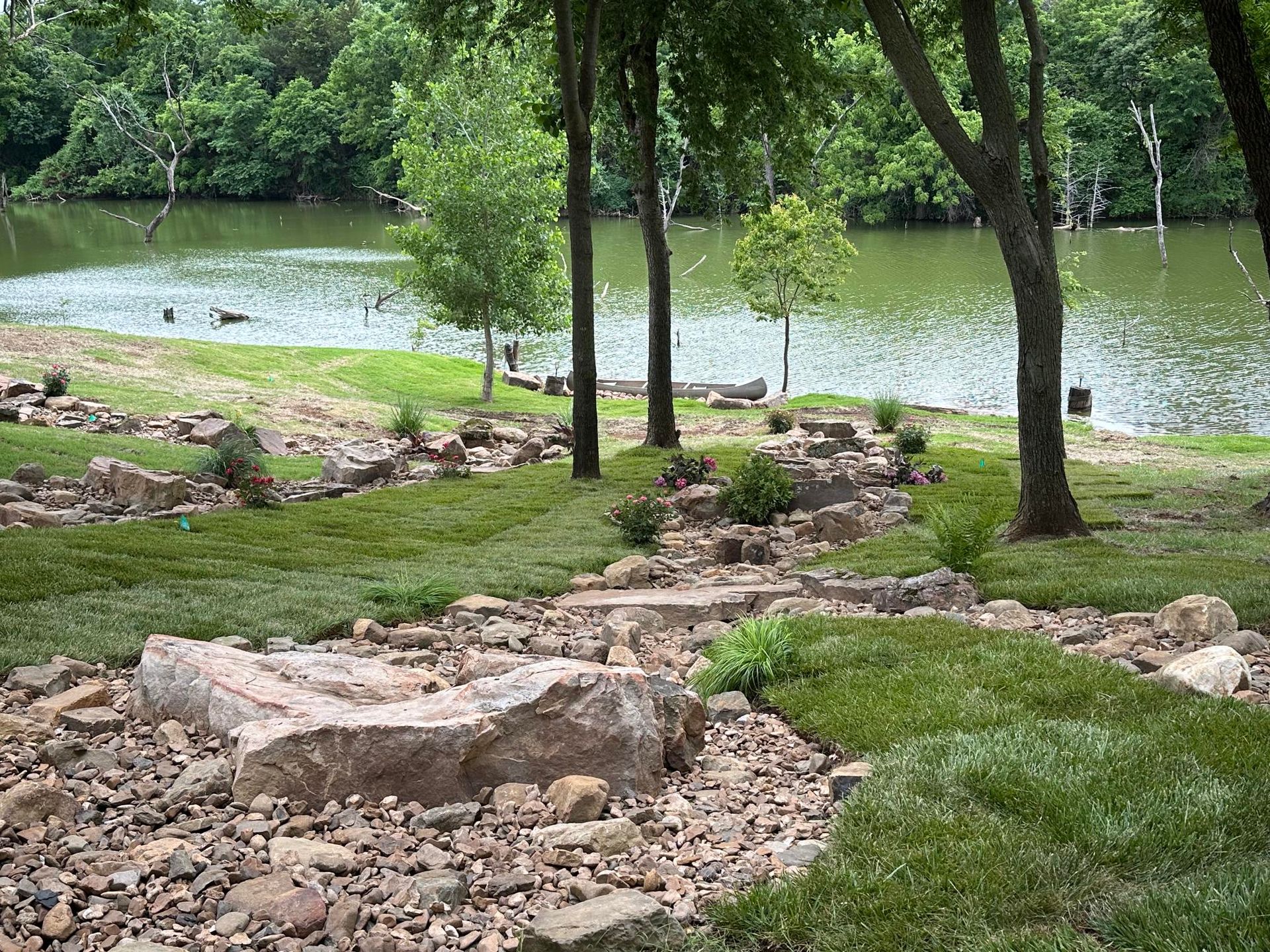 Rocky path through a grassy park leading to a calm lake with trees along the shore