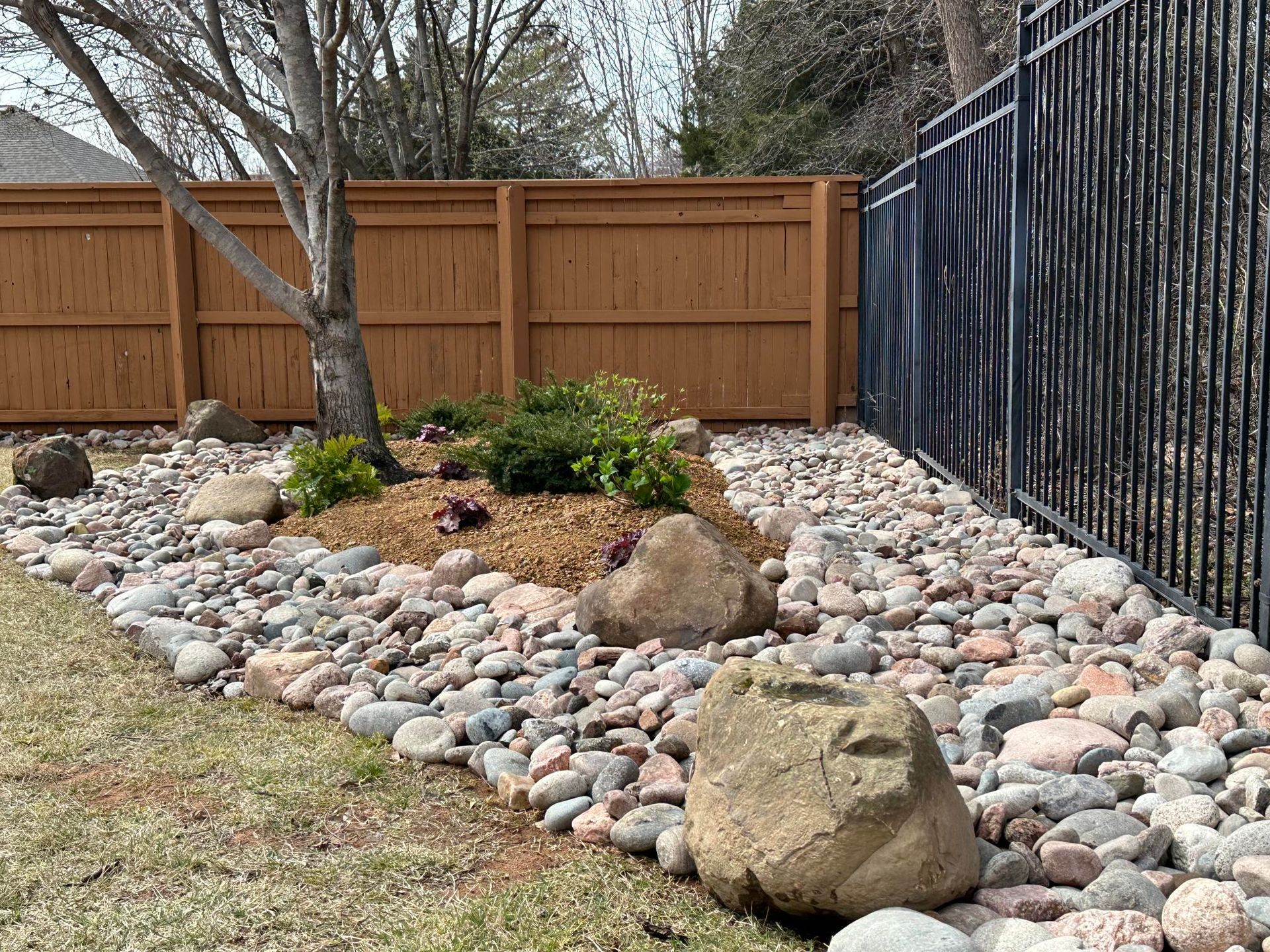 Rock garden with large boulders, small shrubs, and river rocks beside a wooden fence and metal gate.