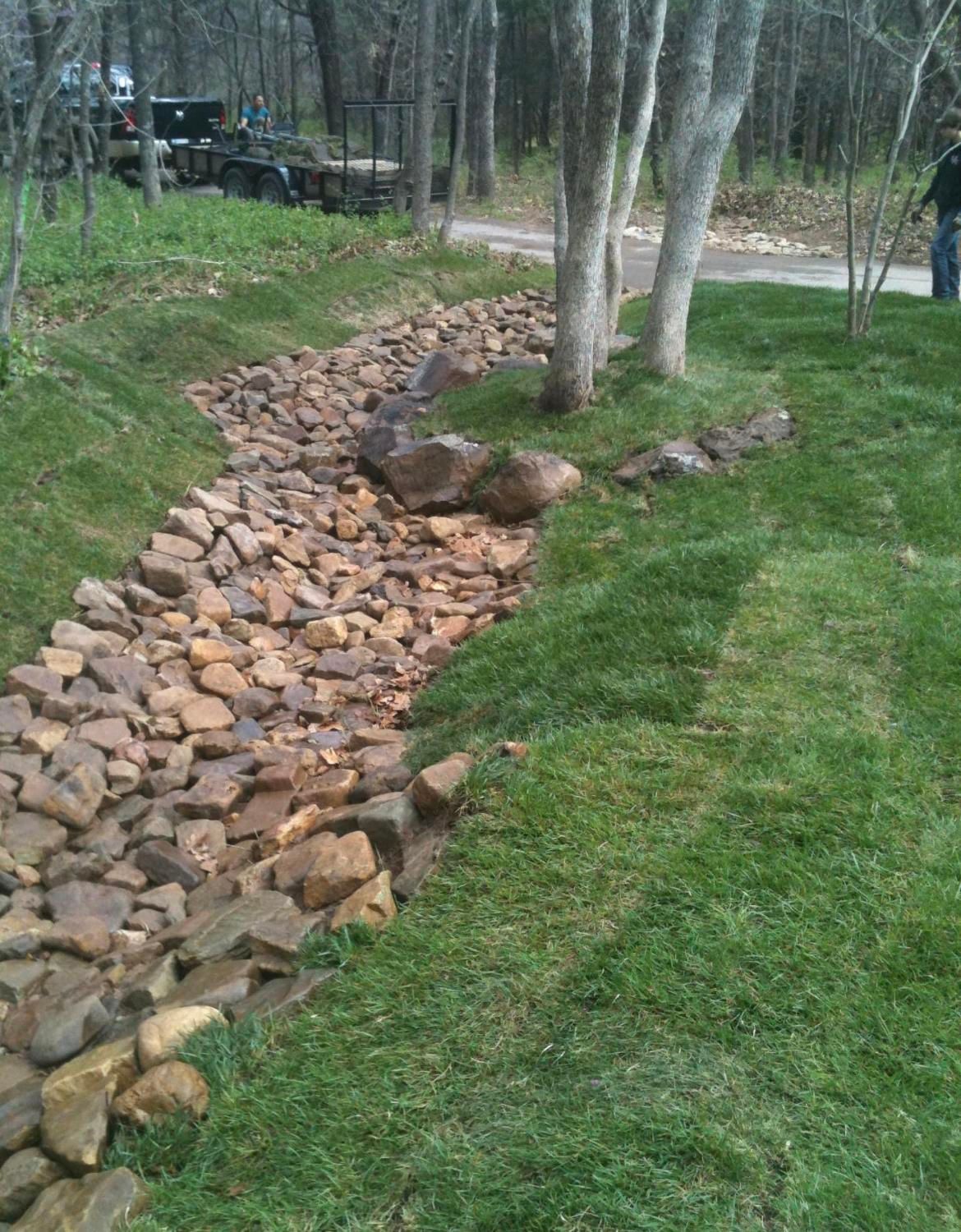 Rock-lined drainage ditch beside a grassy yard with trees and a dirt road.