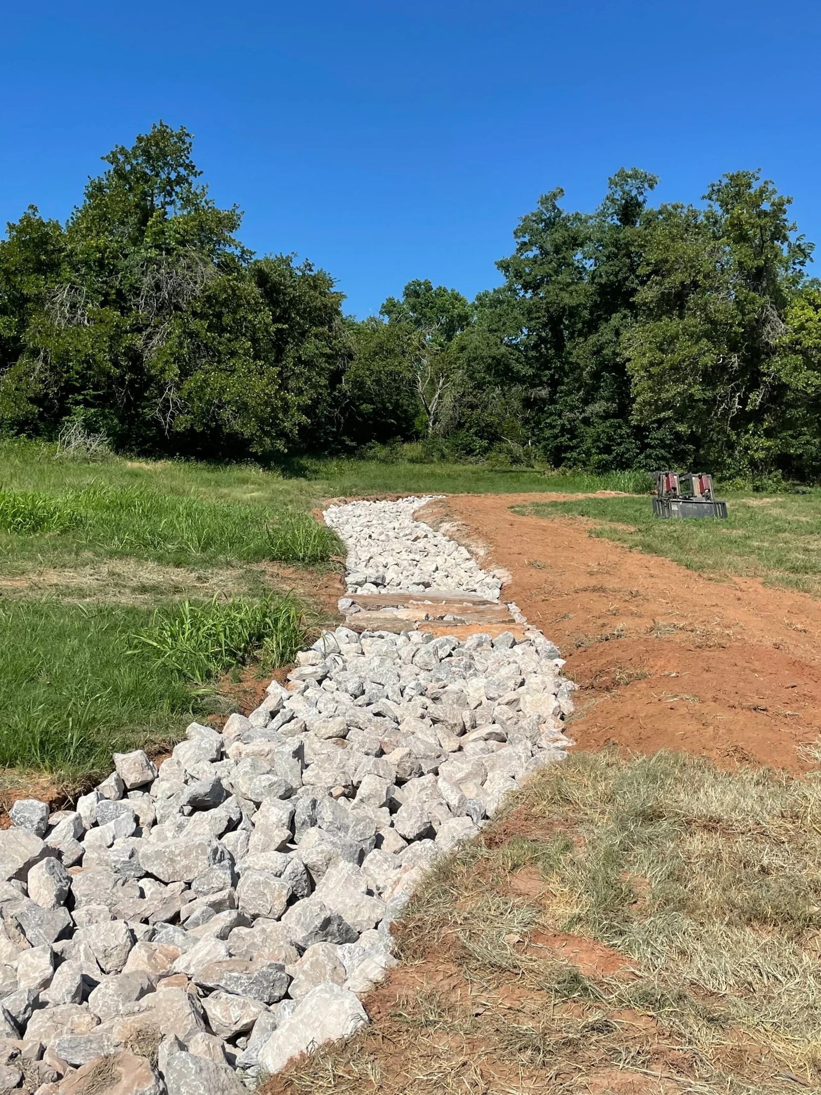 Rock-lined drainage path cutting through a grassy field under a clear blue sky.