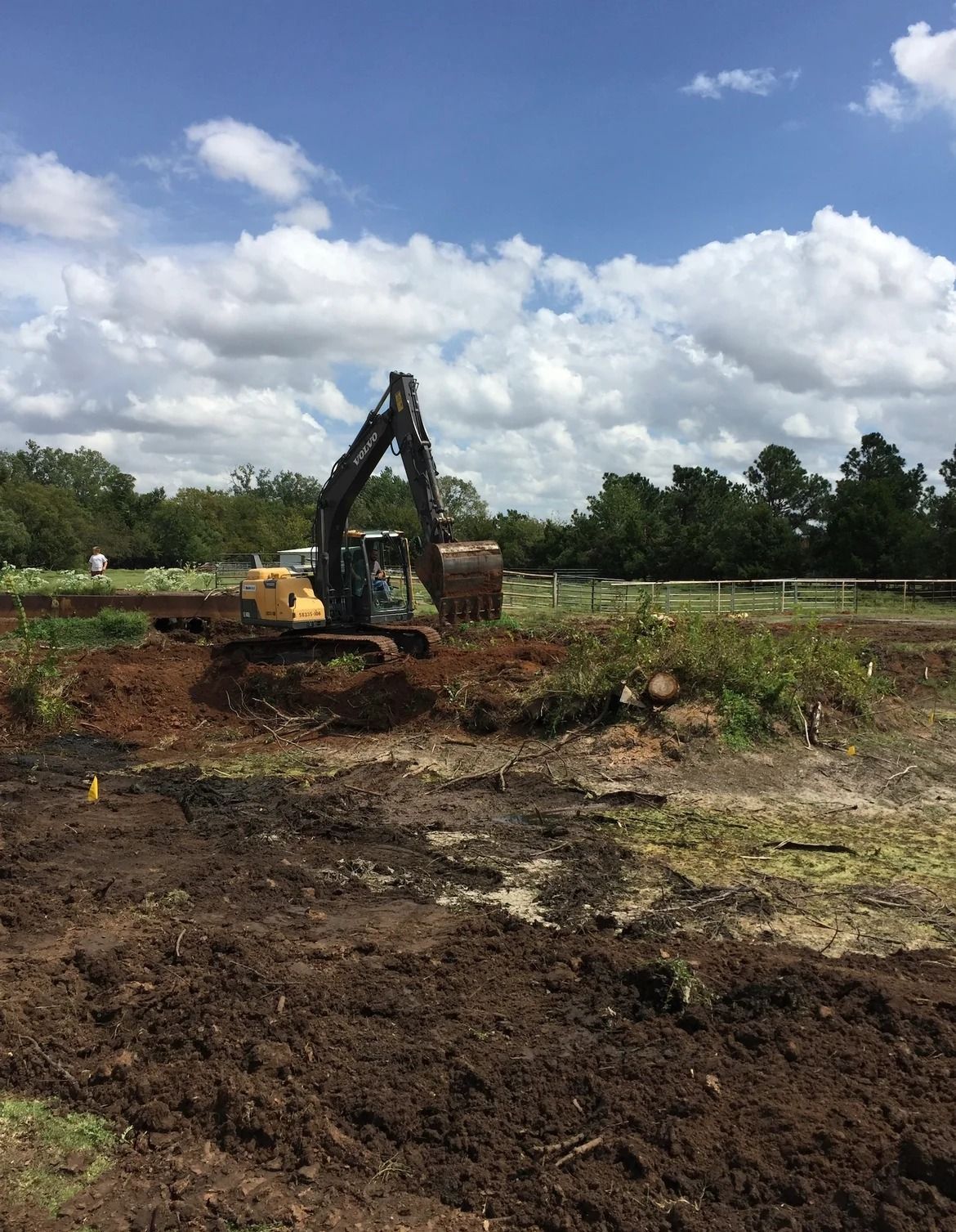 Excavator digging dark soil in a muddy field under a blue sky with clouds.