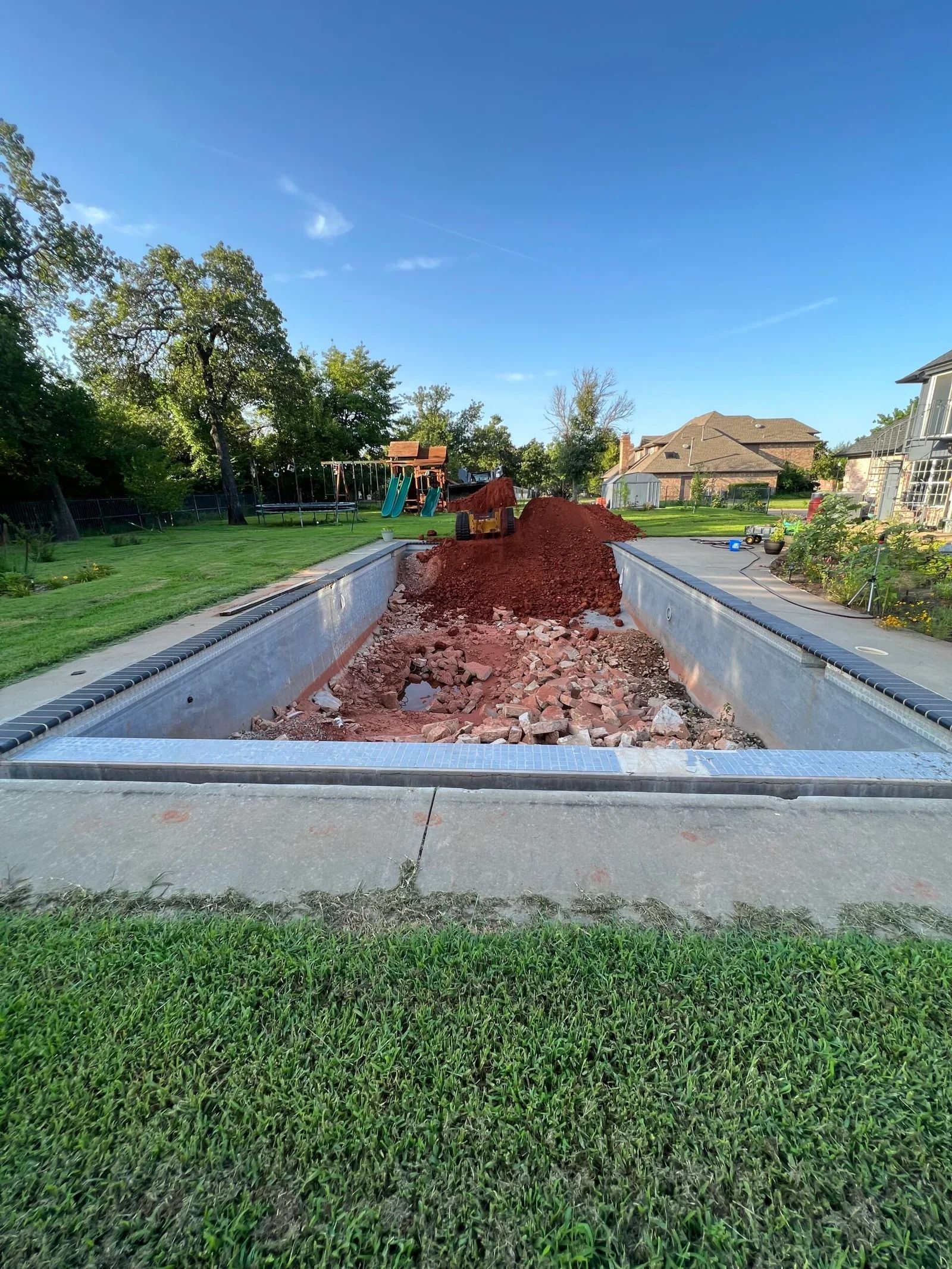 In-ground pool under construction with red dirt, concrete walls, and grassy yard under a clear blue sky