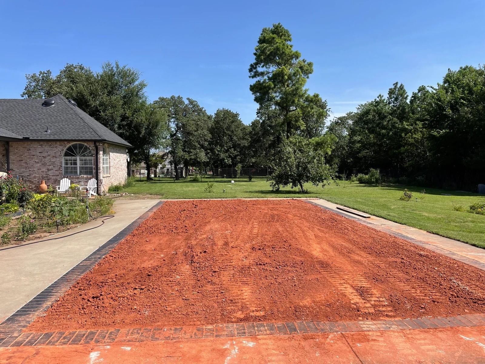 Rectangular red clay tennis court beside a stone house, surrounded by grass and trees under a blue sky
