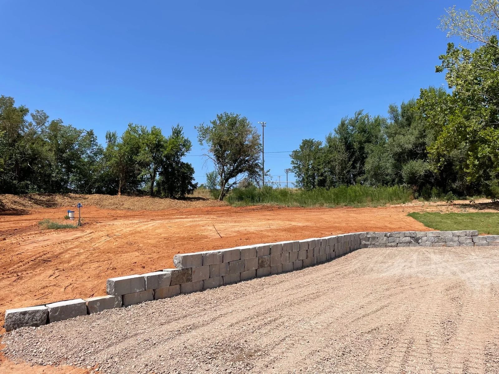 Gravel lot beside a low cinderblock retaining wall and freshly graded dirt under a clear blue sky.