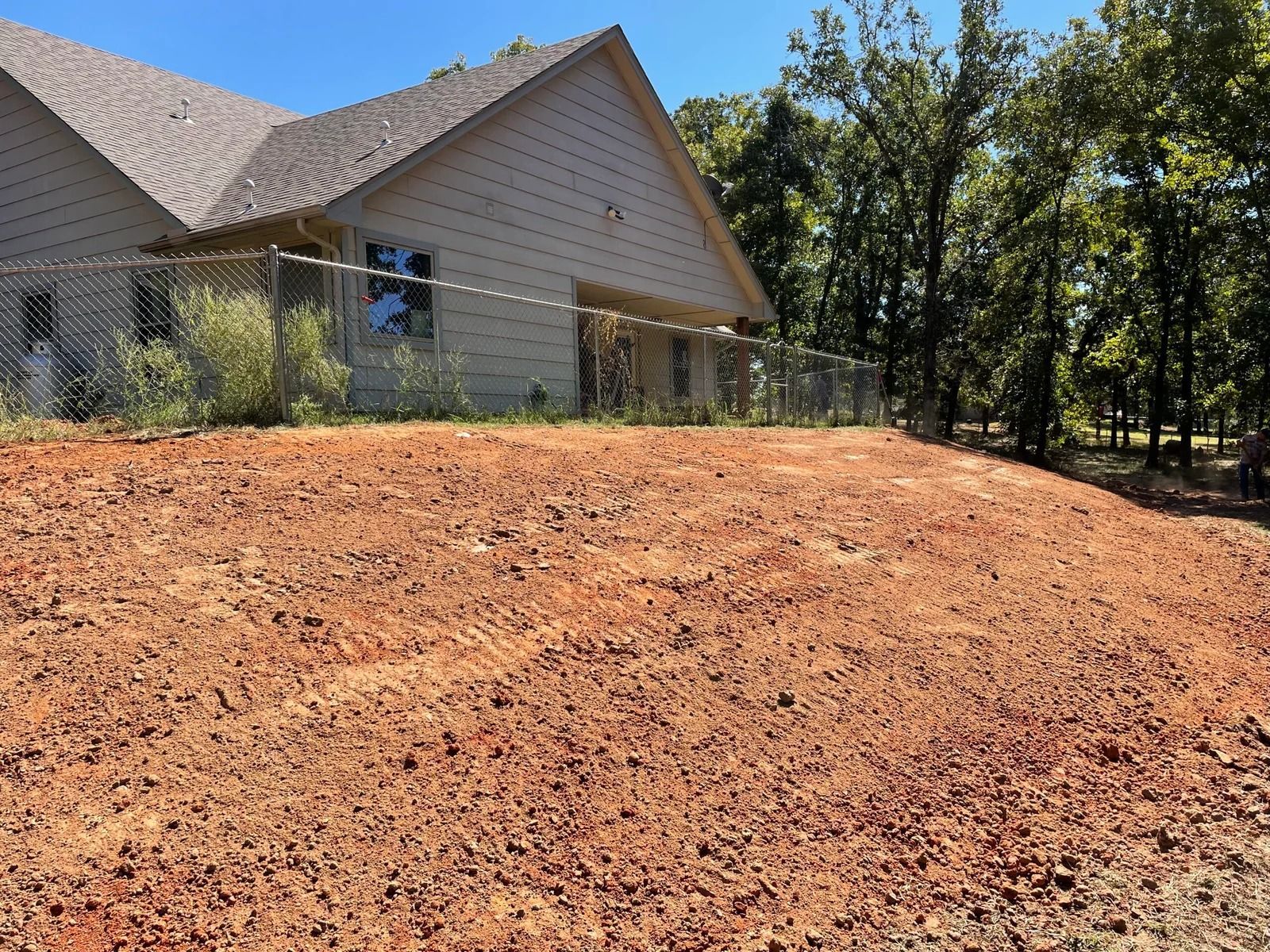 House with beige siding beside a freshly graded dirt slope and trees under a clear blue sky