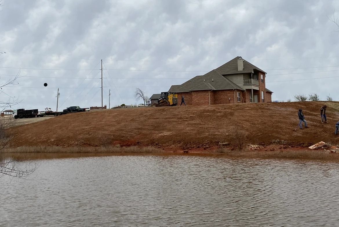 Lakeside house on a brown embankment under a cloudy sky, with water in the foreground.