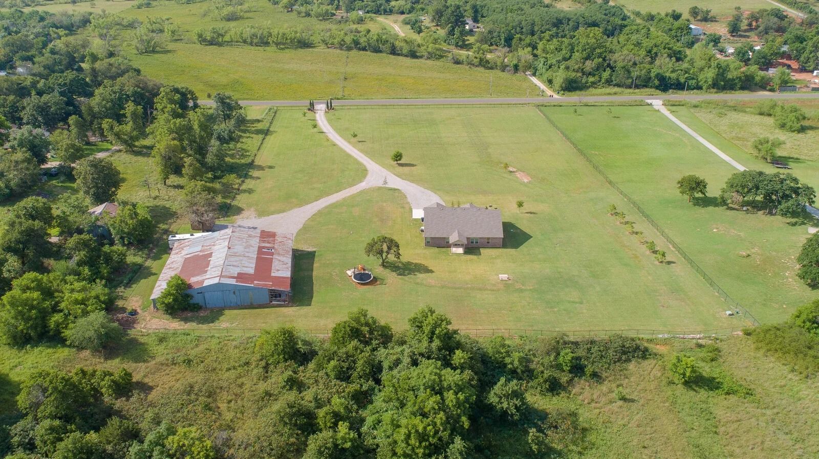 Aerial view of a rural farmstead with a barn, house, fields, and a winding driveway surrounded by trees.