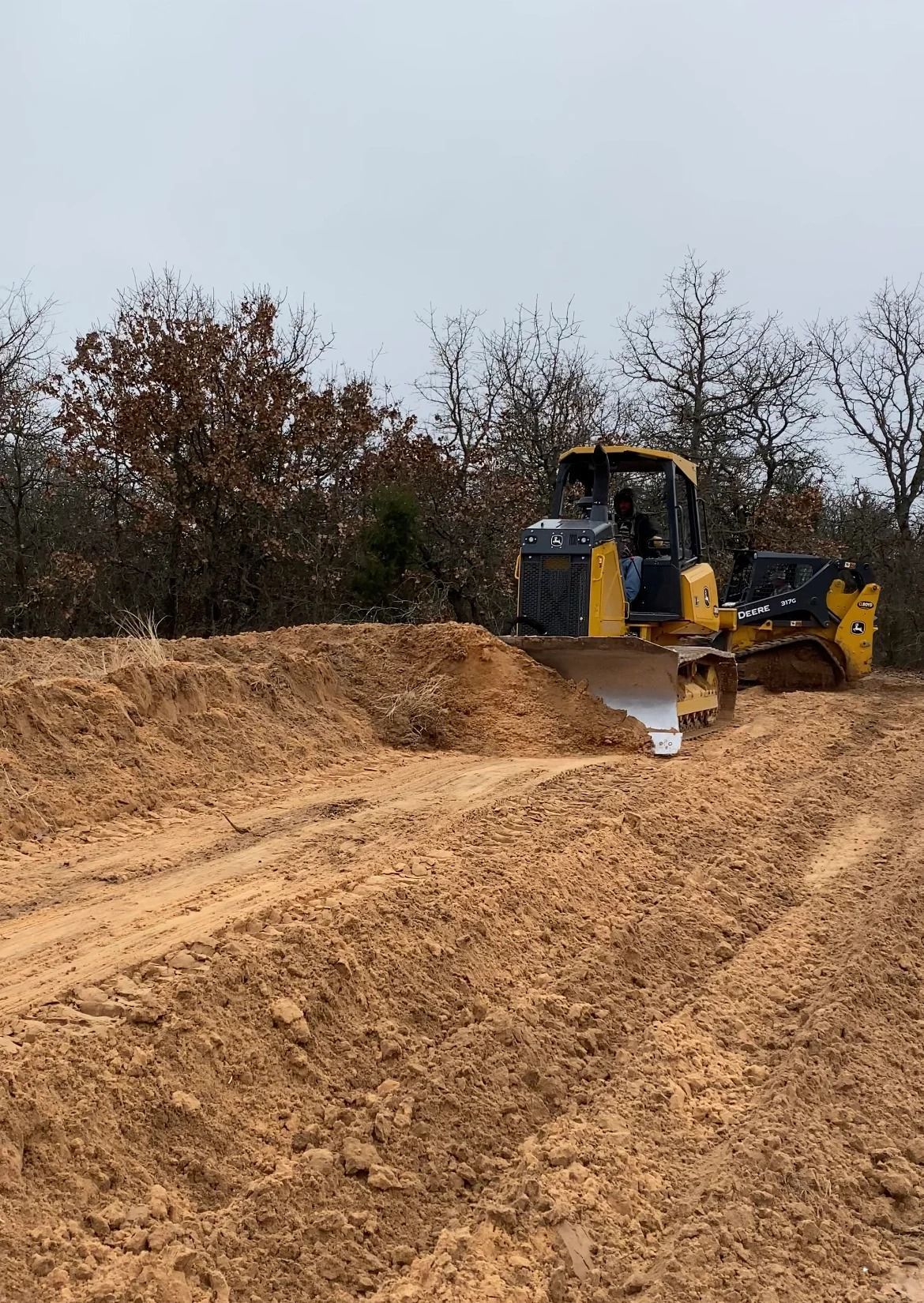 Yellow excavator leveling a dirt road under a gray sky, with trees in the background