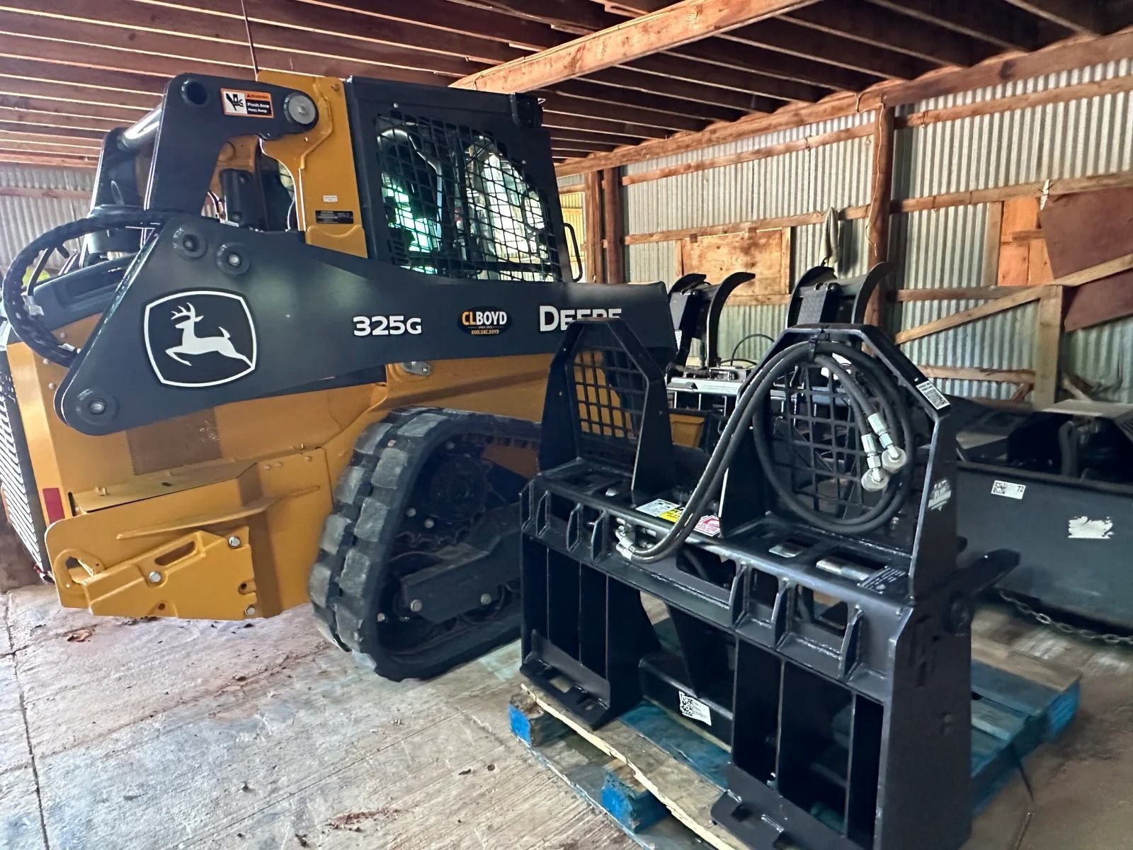 Yellow John Deere 325G compact track loader in a barn, beside a black metal attachment frame.