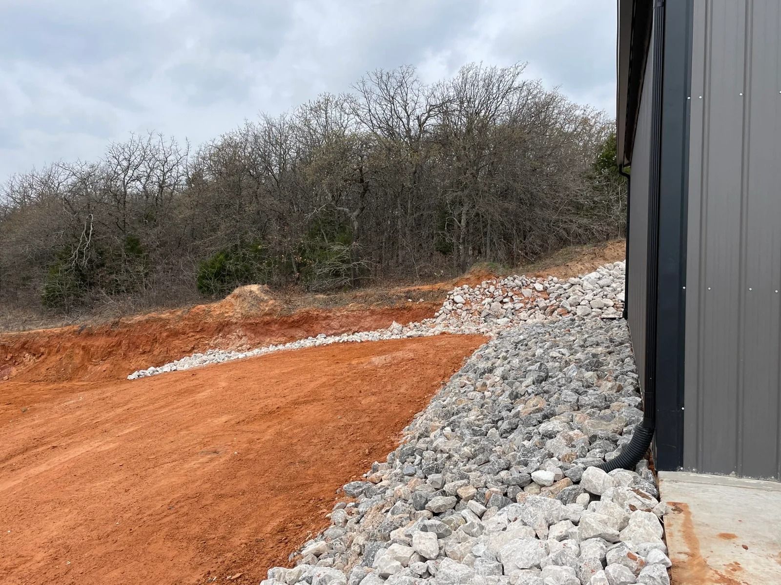 Red dirt path beside gray building, lined with rocks and brush on a cloudy day