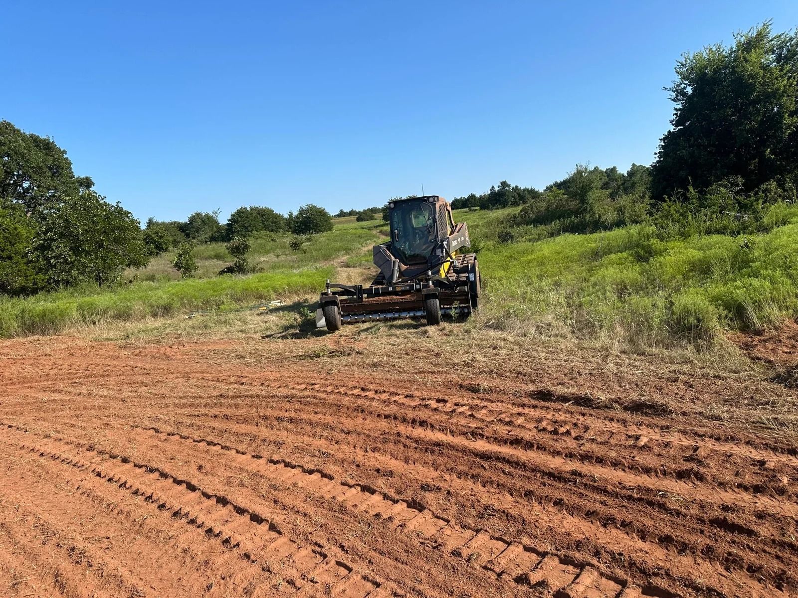 Bulldozer grading a dirt field under a clear blue sky, with green trees in the background