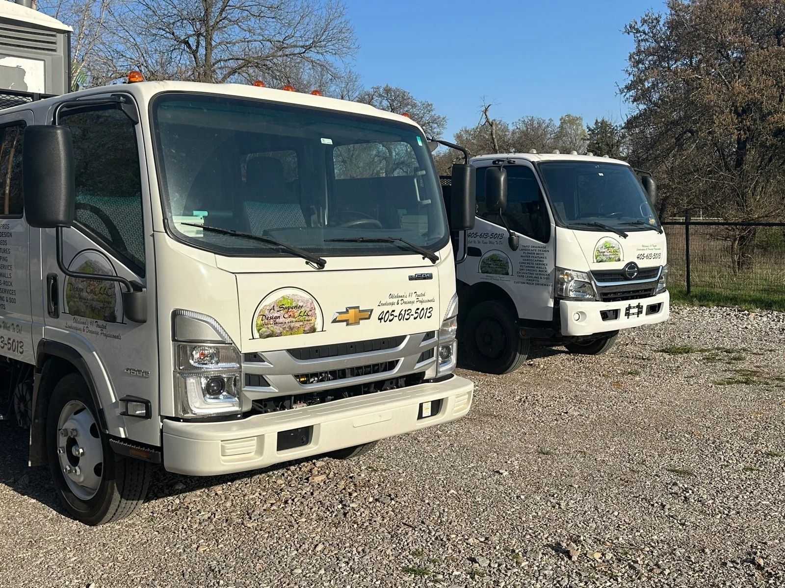 Two white box trucks parked on gravel in front of a building under a clear blue sky