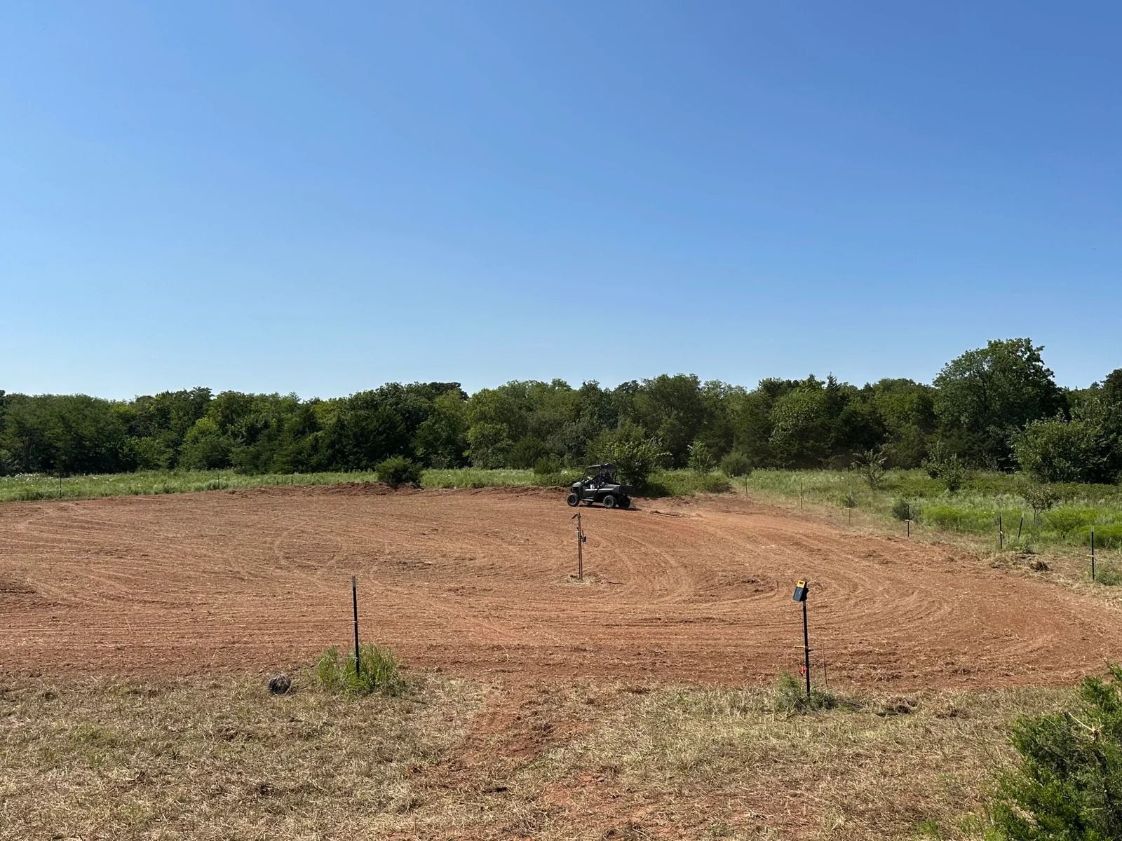Dry, cleared field with a small vehicle in the distance under a blue sky, bordered by trees.