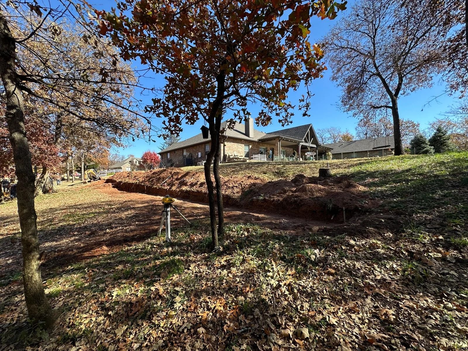 Autumn yard with a house on a hill, bare trees, and a reddish dirt slope under a blue sky
