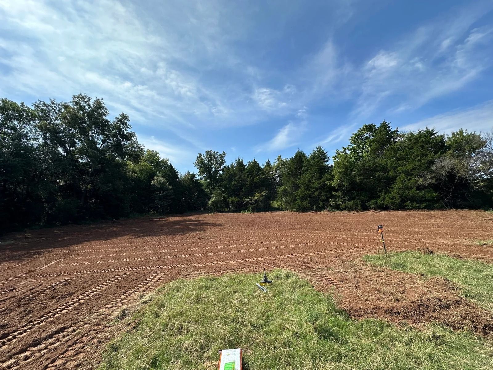Cleared field with bare soil and grass under a blue sky, bordered by trees and shrubs
