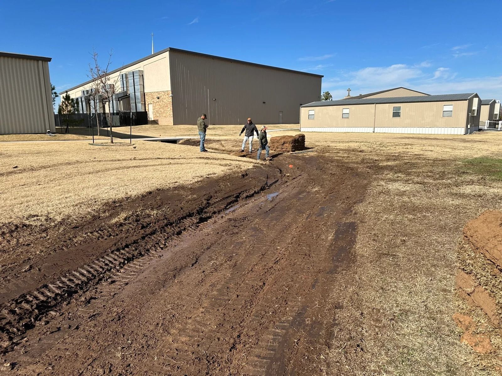 People working beside a muddy dirt path between tan buildings under a clear blue sky.