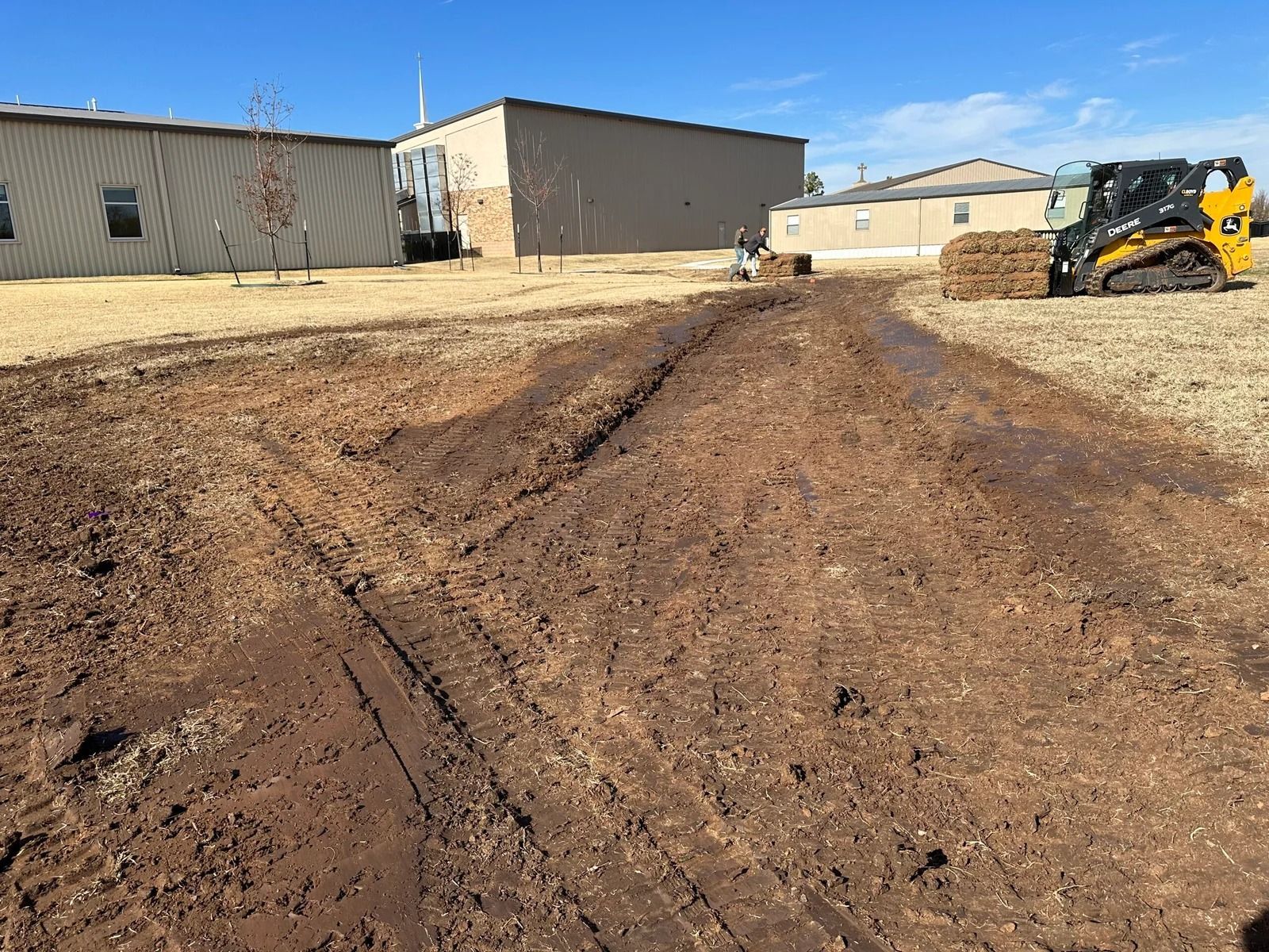 Mud track in a construction yard beside buildings and a yellow skid steer.