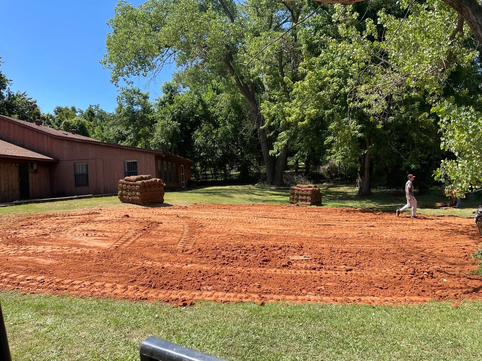 Freshly graded red dirt yard beside a brown building, with stacked pavers and a person near trees