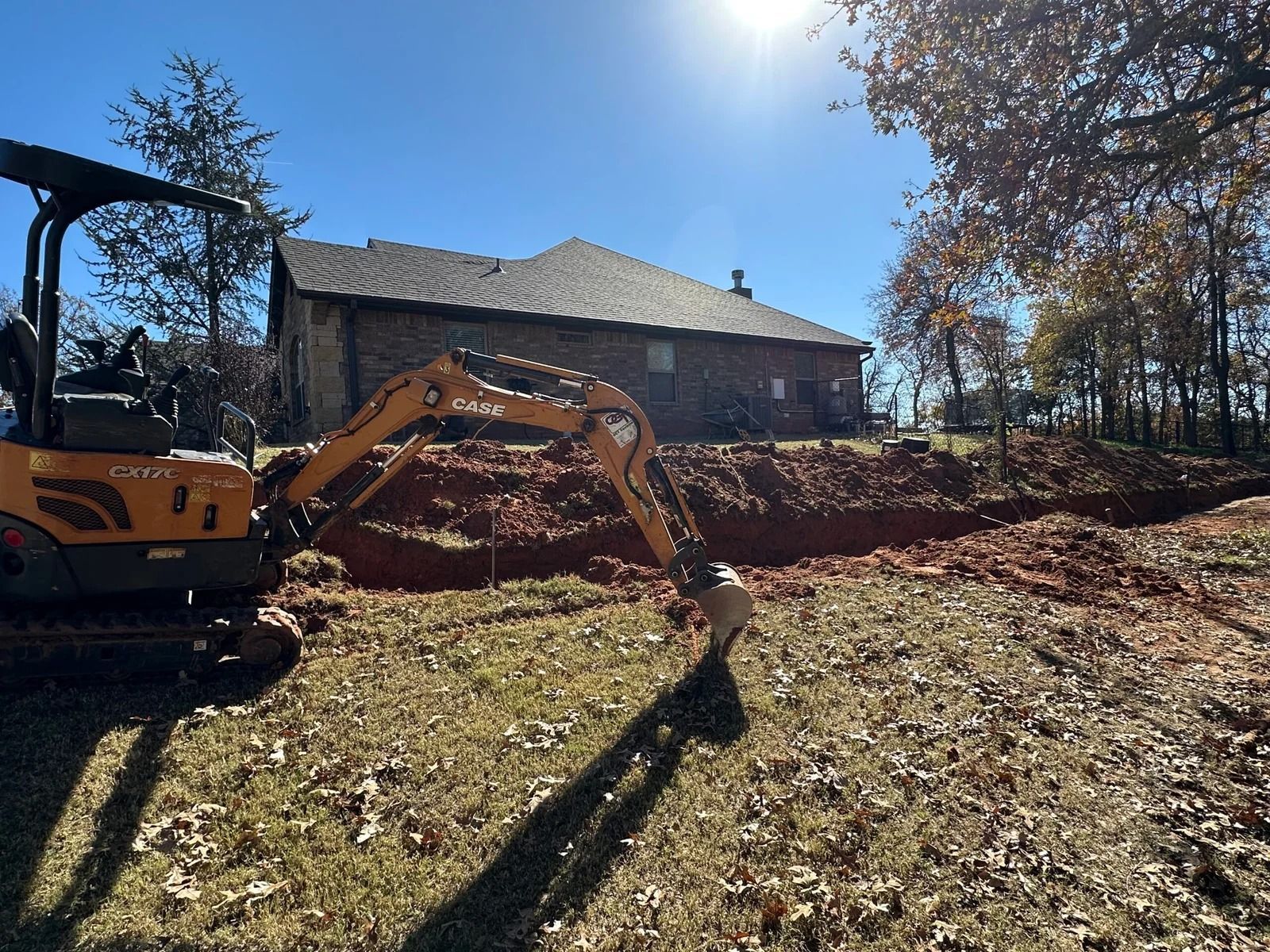 Excavator digging in a leaf-covered yard beside a house under a bright blue sky