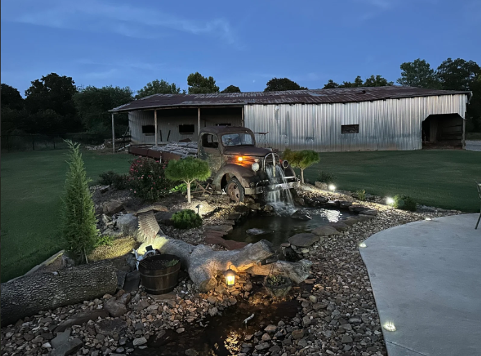 Backyard pond with fountain and garden lights beside a house at dusk