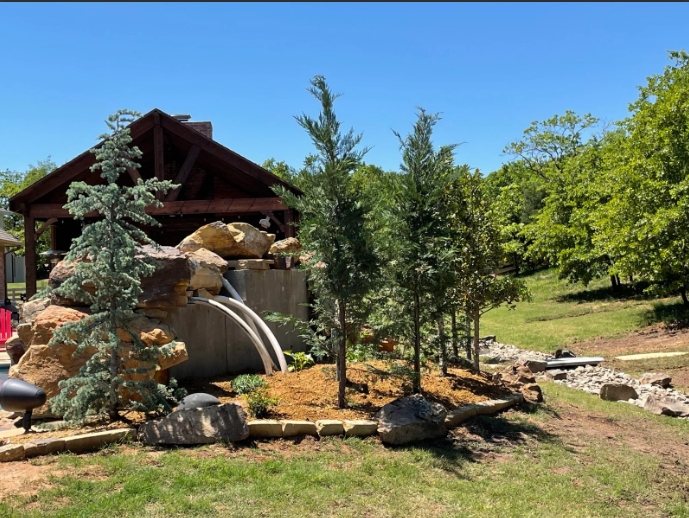 Rustic backyard waterfall garden with rocks, trees, and a wooden cabin under a blue sky