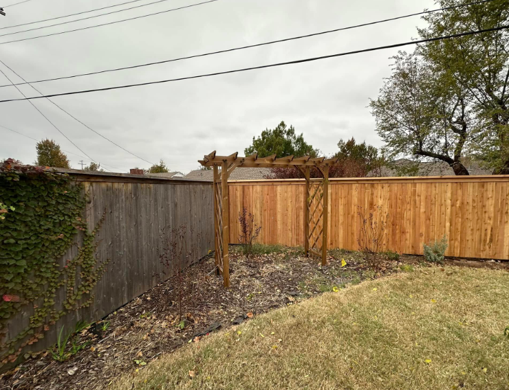 Backyard corner with wooden privacy fence, trellis post, and grassy lawn under cloudy sky