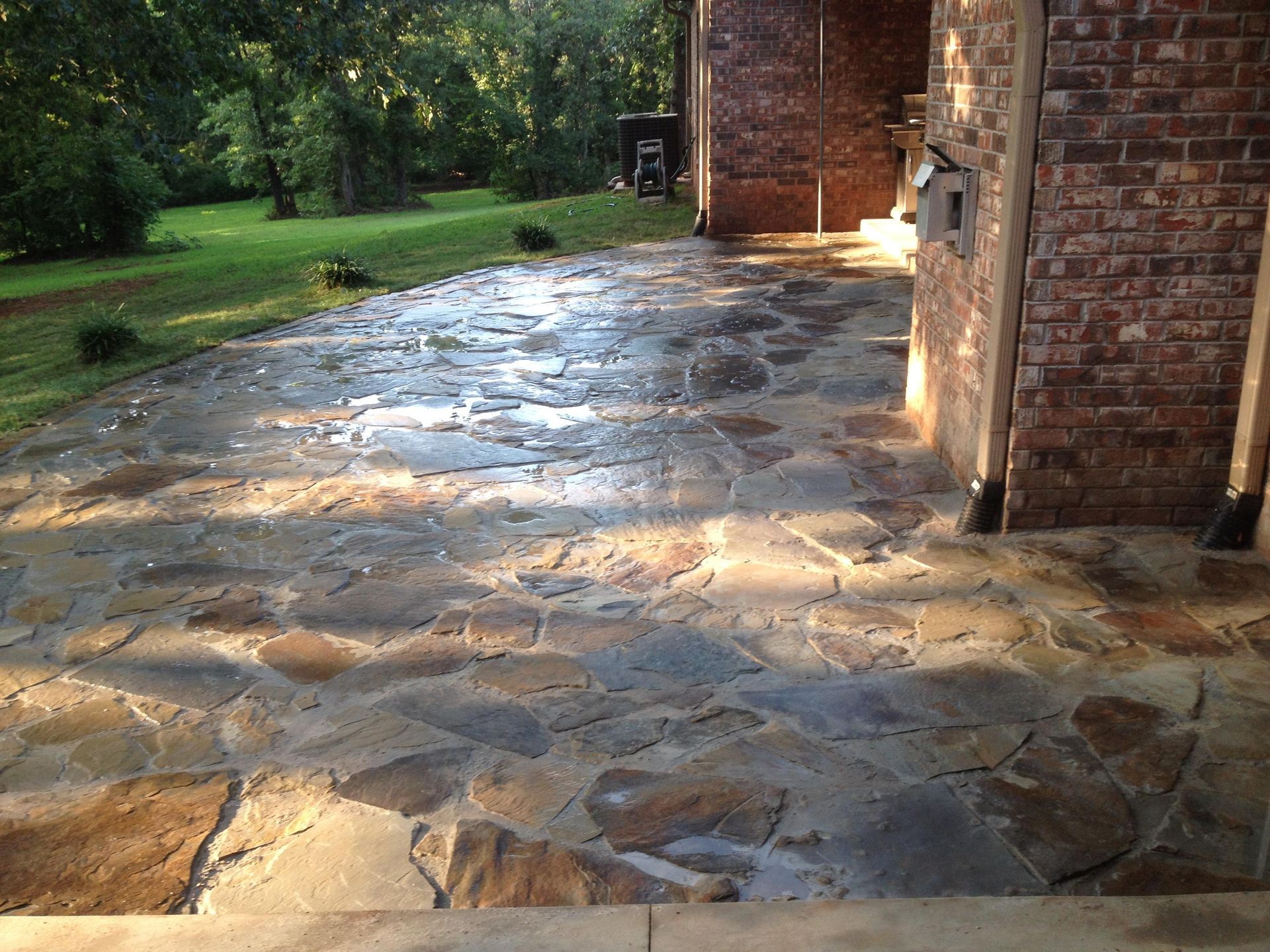 Stone patio beside a brick house, sunlit and wet, with a grassy yard in the background