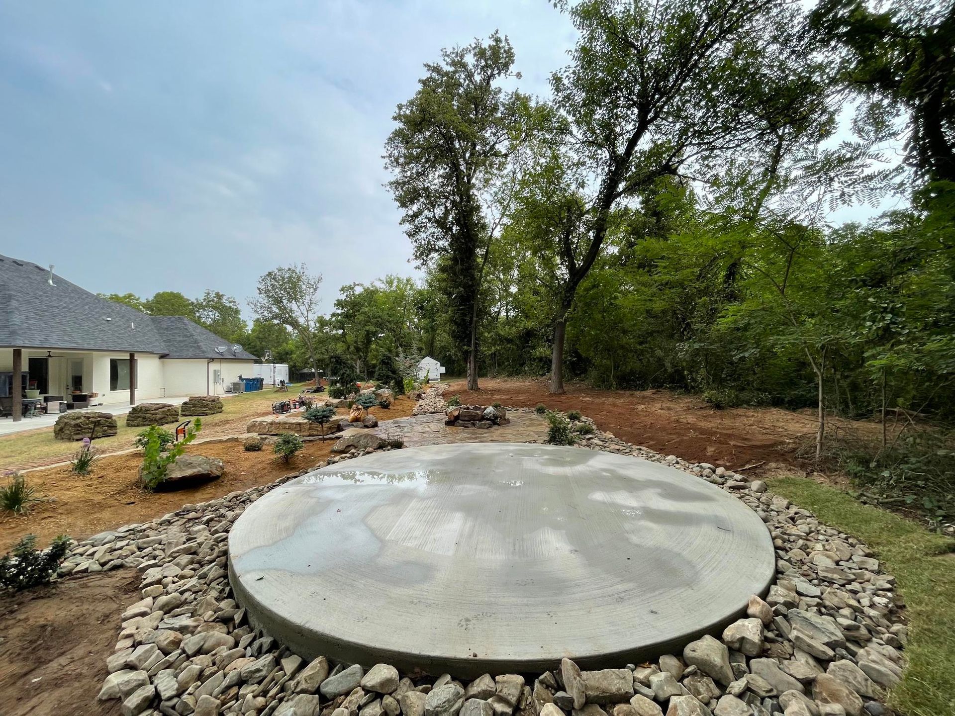 Circular concrete pad in a landscaped backyard with rocks, trees, and a house in the background