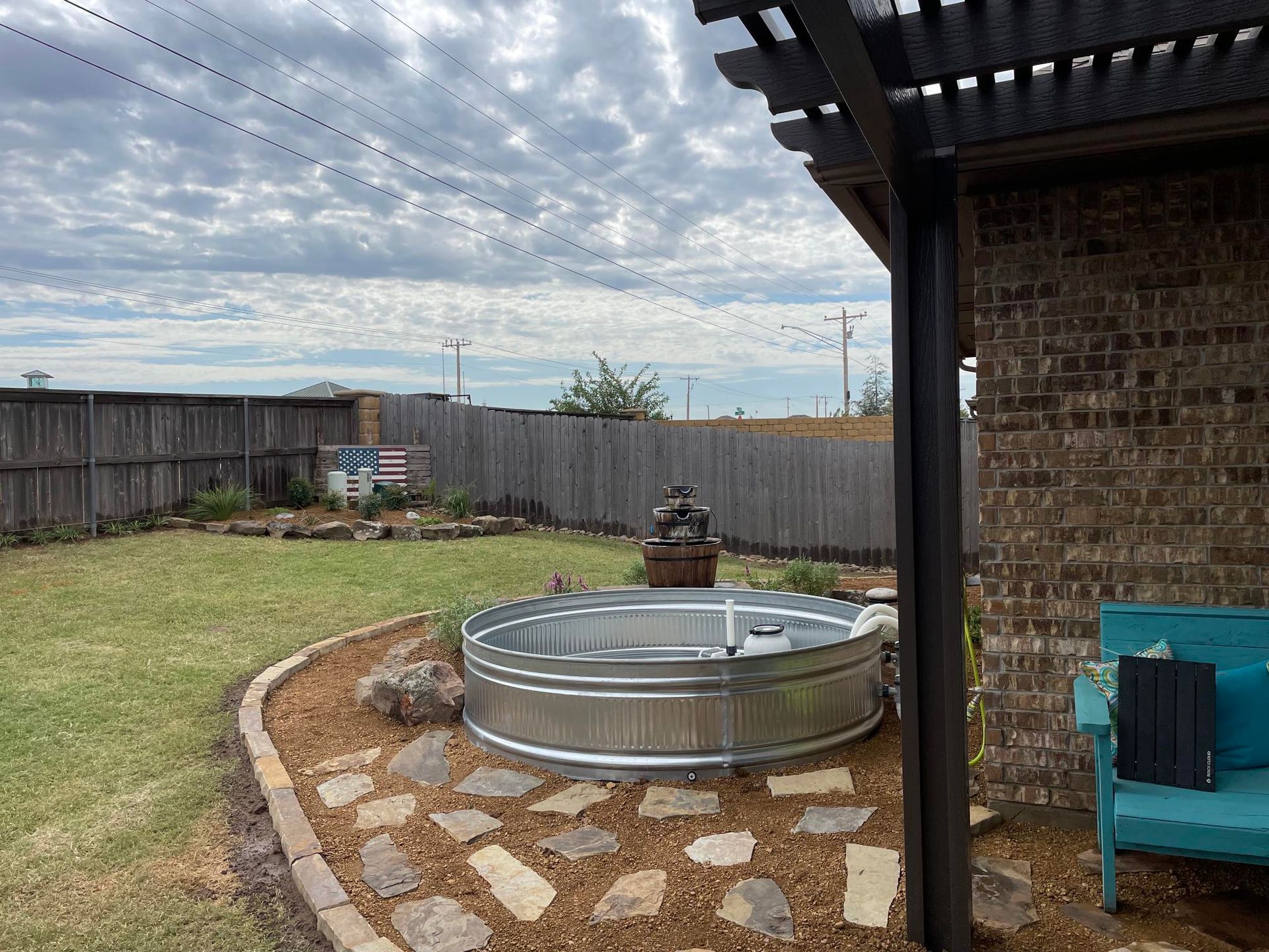 Backyard with a metal above-ground pool, stone patio, grass, wooden fence, and cloudy sky