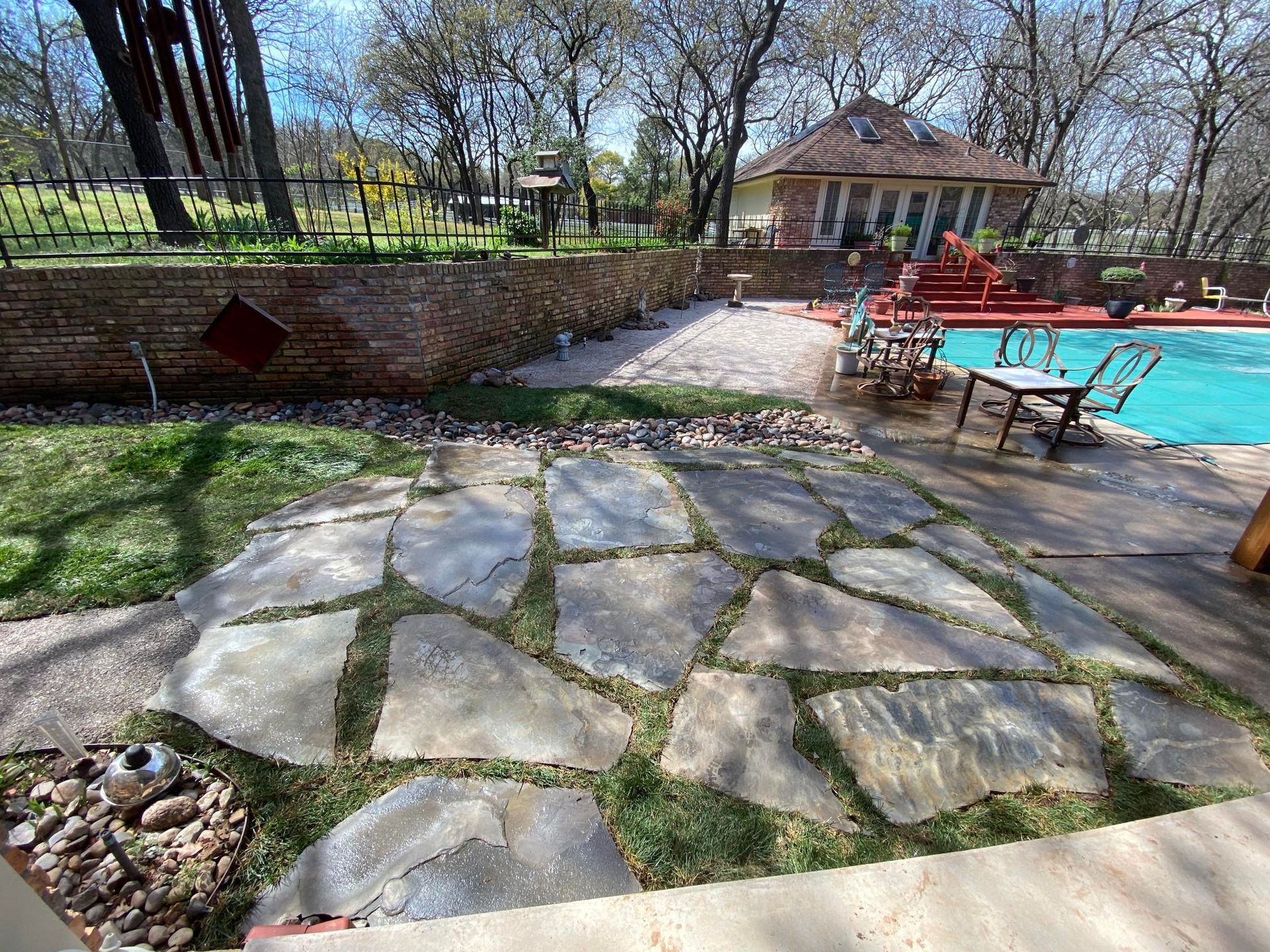 Stone patio path beside a turquoise pool, with red chairs, brick wall, and gazebo in a sunny yard