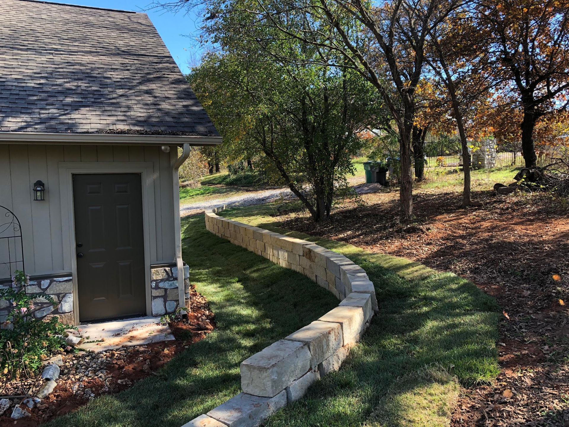 Curved stone border beside a shed, leading through a grassy yard with trees and autumn leaves.