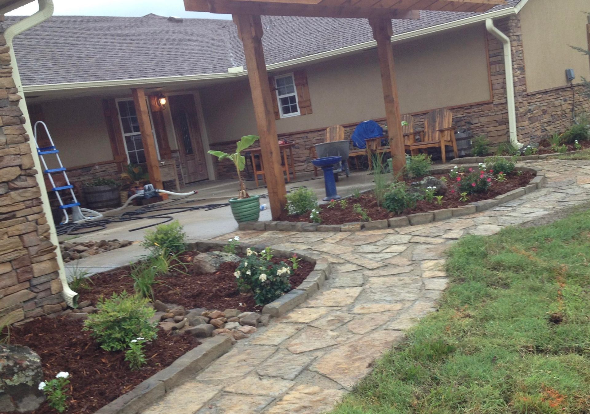 Stone walkway through a landscaped front yard leading to a covered porch with flower beds and a ladder nearby