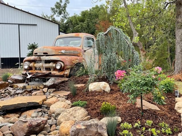 Rusty vintage truck in a landscaped garden with rocks, flowers, and a white shed in the background