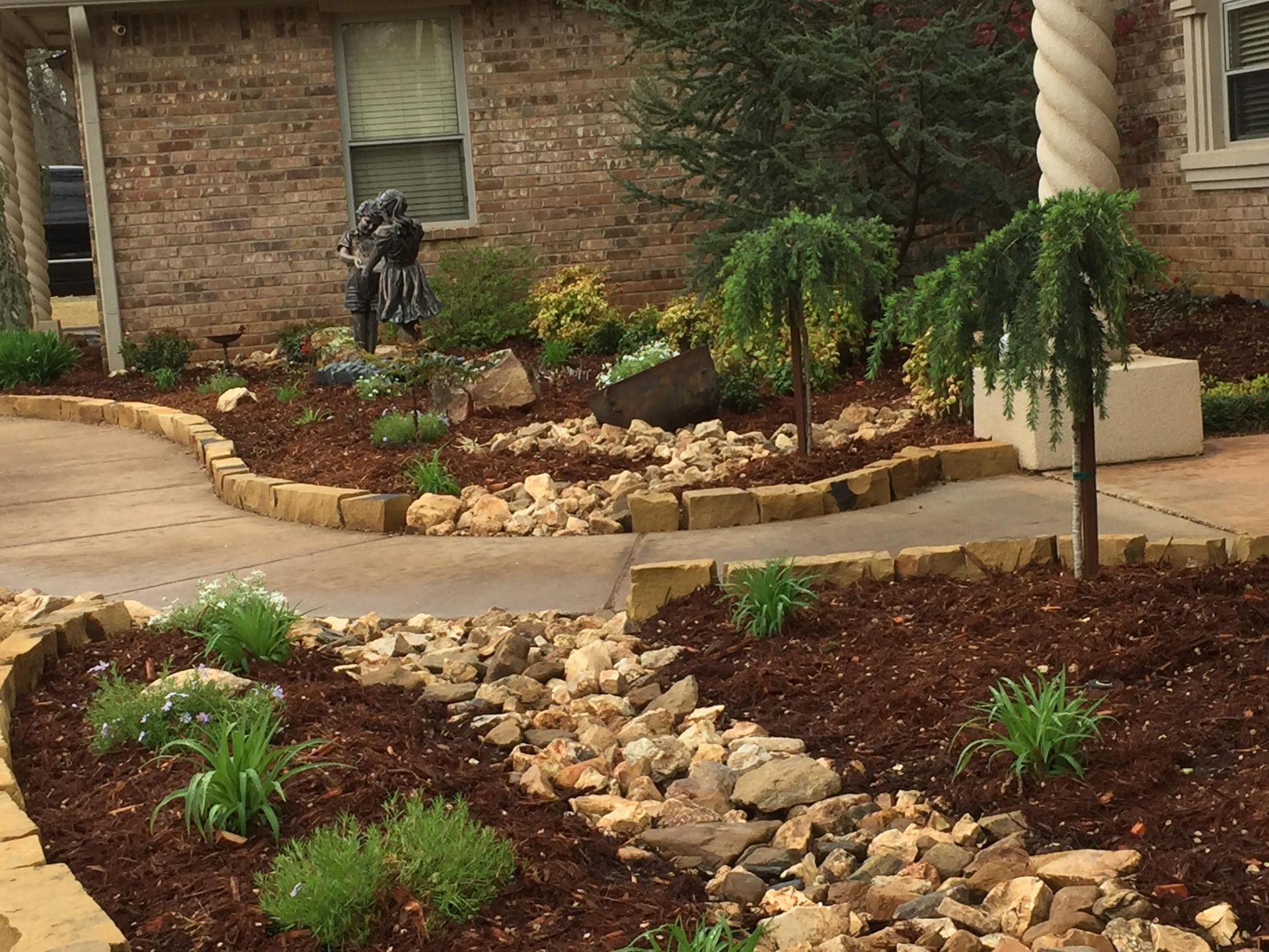 Front yard landscaping with stone border, mulch beds, small shrubs, and a rock-lined dry creek bed.